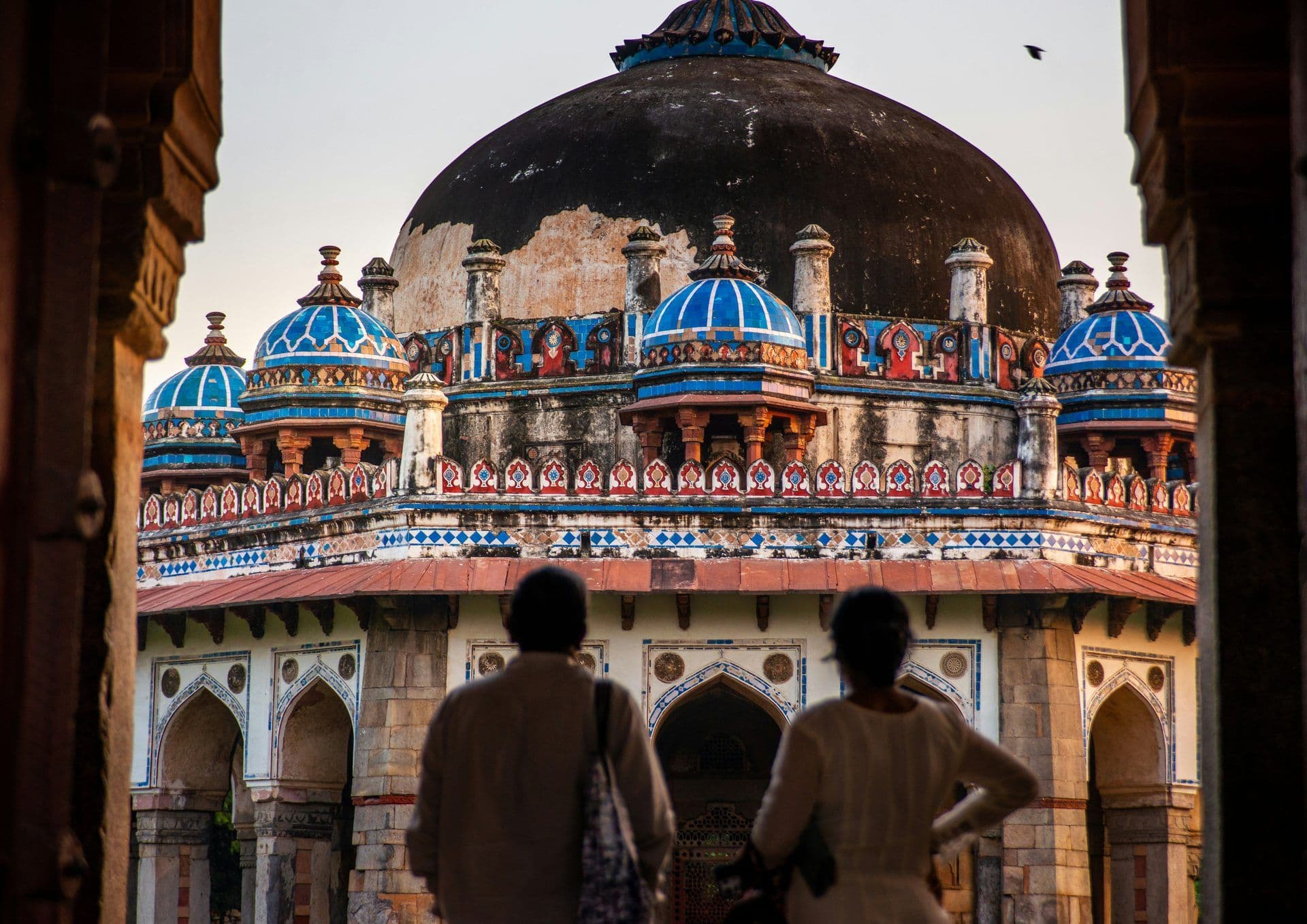 Two people view a historic Indian monument with a large dome and blue smaller domes through an archway in