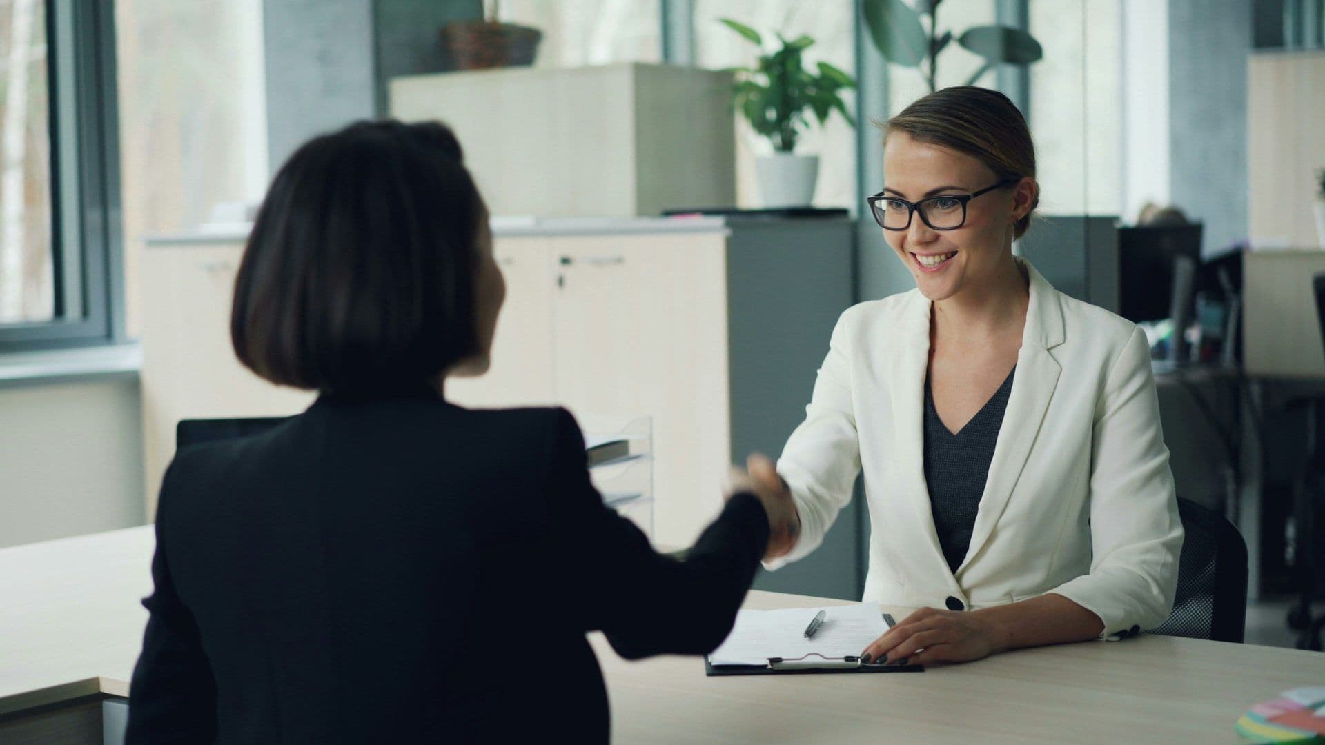 Smiling businesswoman in glasses shakes hands with a colleague across a desk in a bright corporate meeting