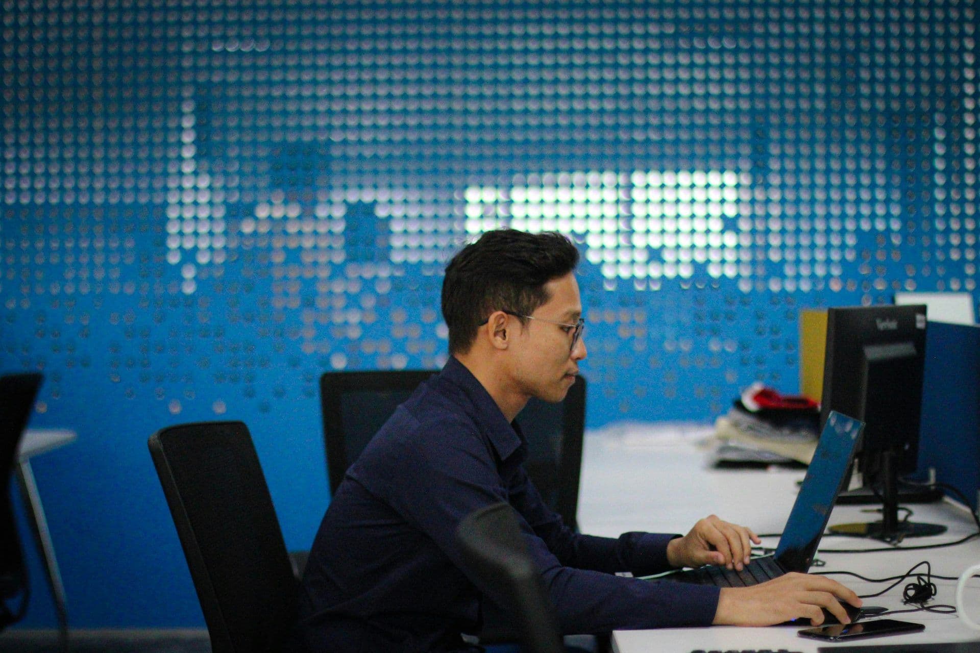 A man works on a laptop at a desk in a modern office or meeting room, with a blue patterned wall.