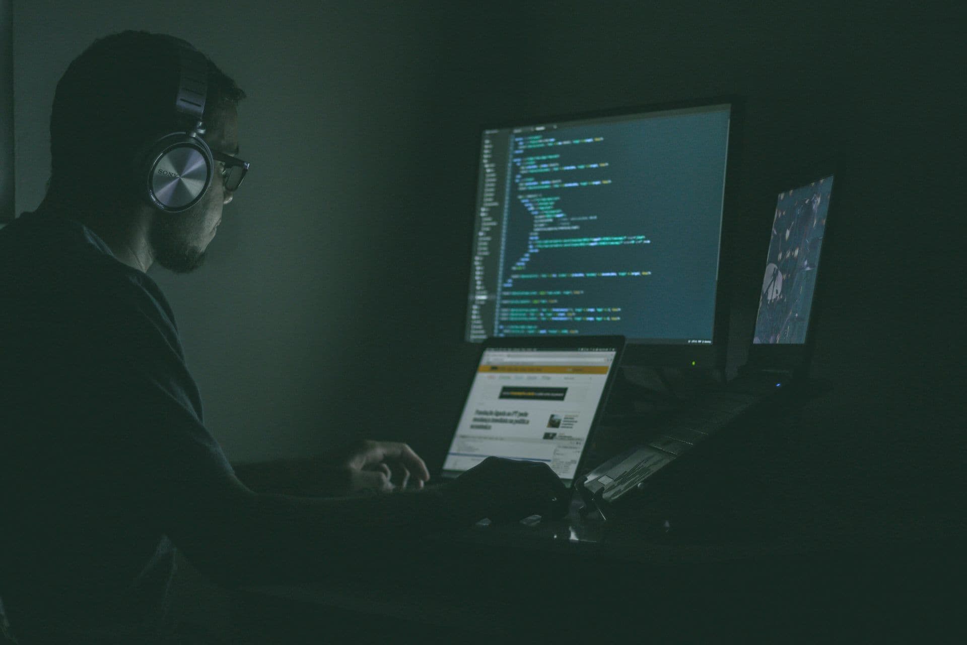 A person with headphones and glasses codes on multiple computer screens in a dark office environment.