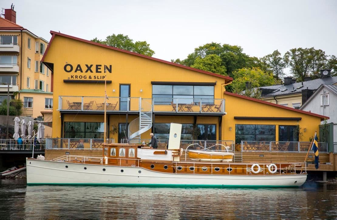 Yellow waterfront event venue Oaxen Krog & Slip with balconies and a classic boat docked in Stockholm.