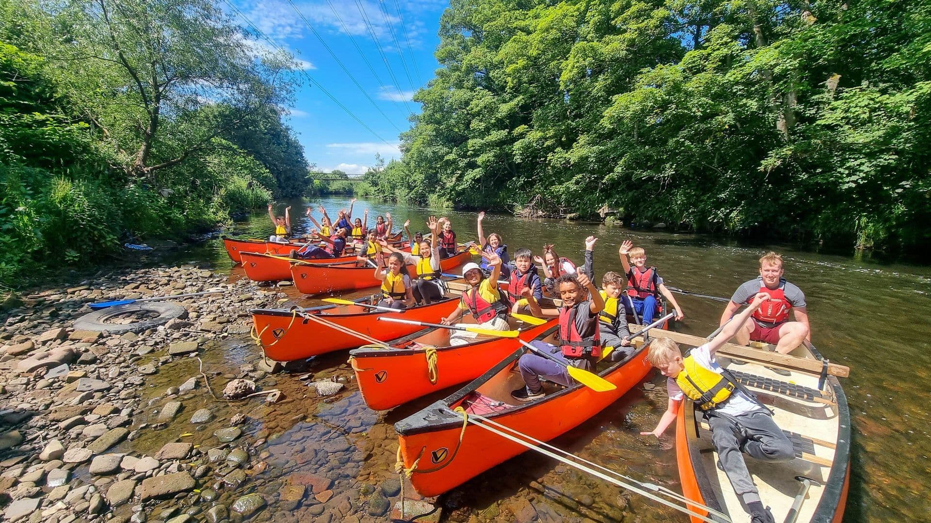 A large group in orange canoes on a river, waving and smiling during an outdoor team building activity.