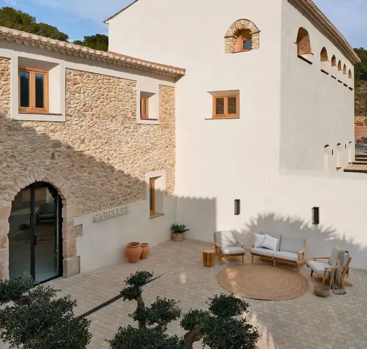 A sunny outdoor courtyard with stone and white buildings, modern seating, and olive trees