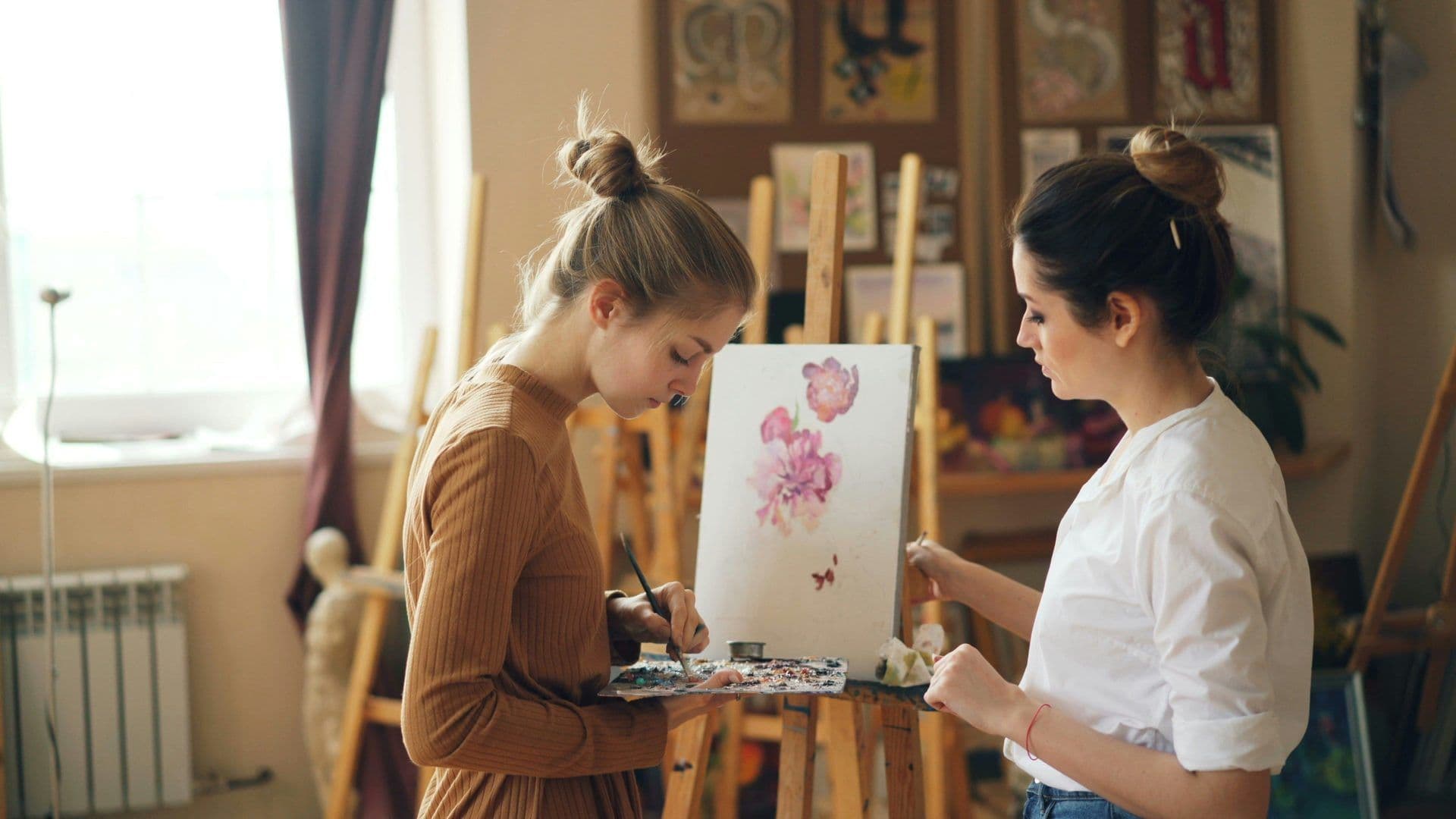 Two women participate in an art workshop, painting flowers on easels in a creative studio space.