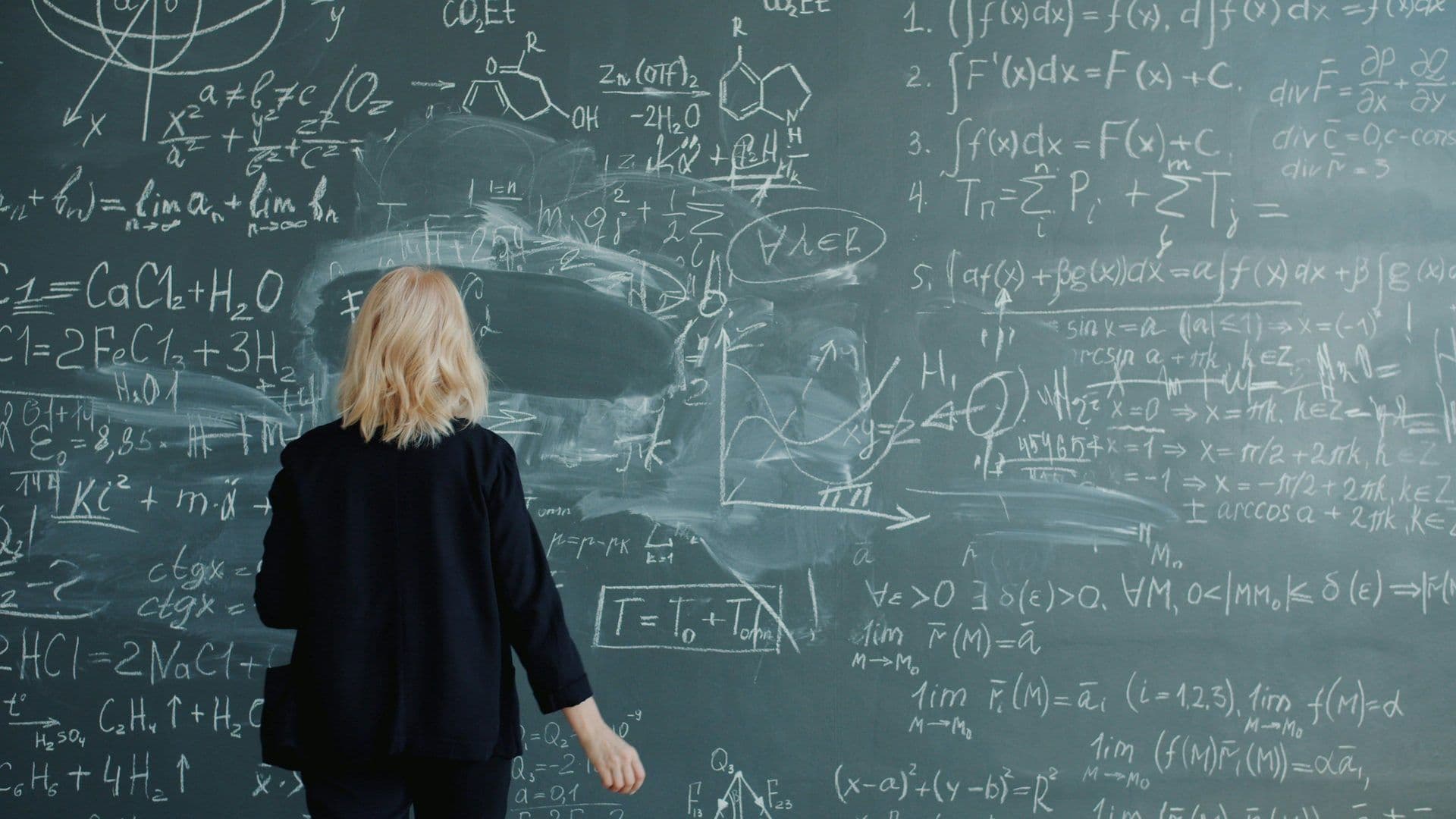 A blonde woman faces a large blackboard filled with intricate mathematical and chemical equations.