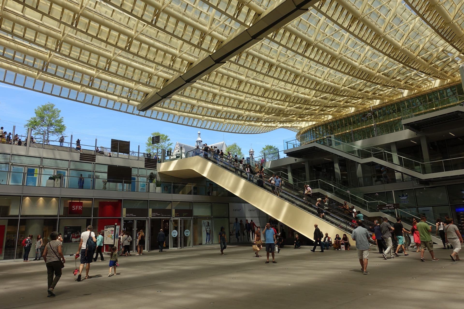 A bustling modern multi-level public event space with a distinctive glass canopy roof and escalators in Paris.