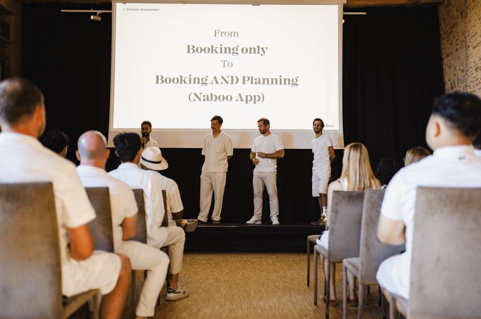Audience watches presenters on stage at a corporate seminar, with 'Naboo App' on the projector screen.