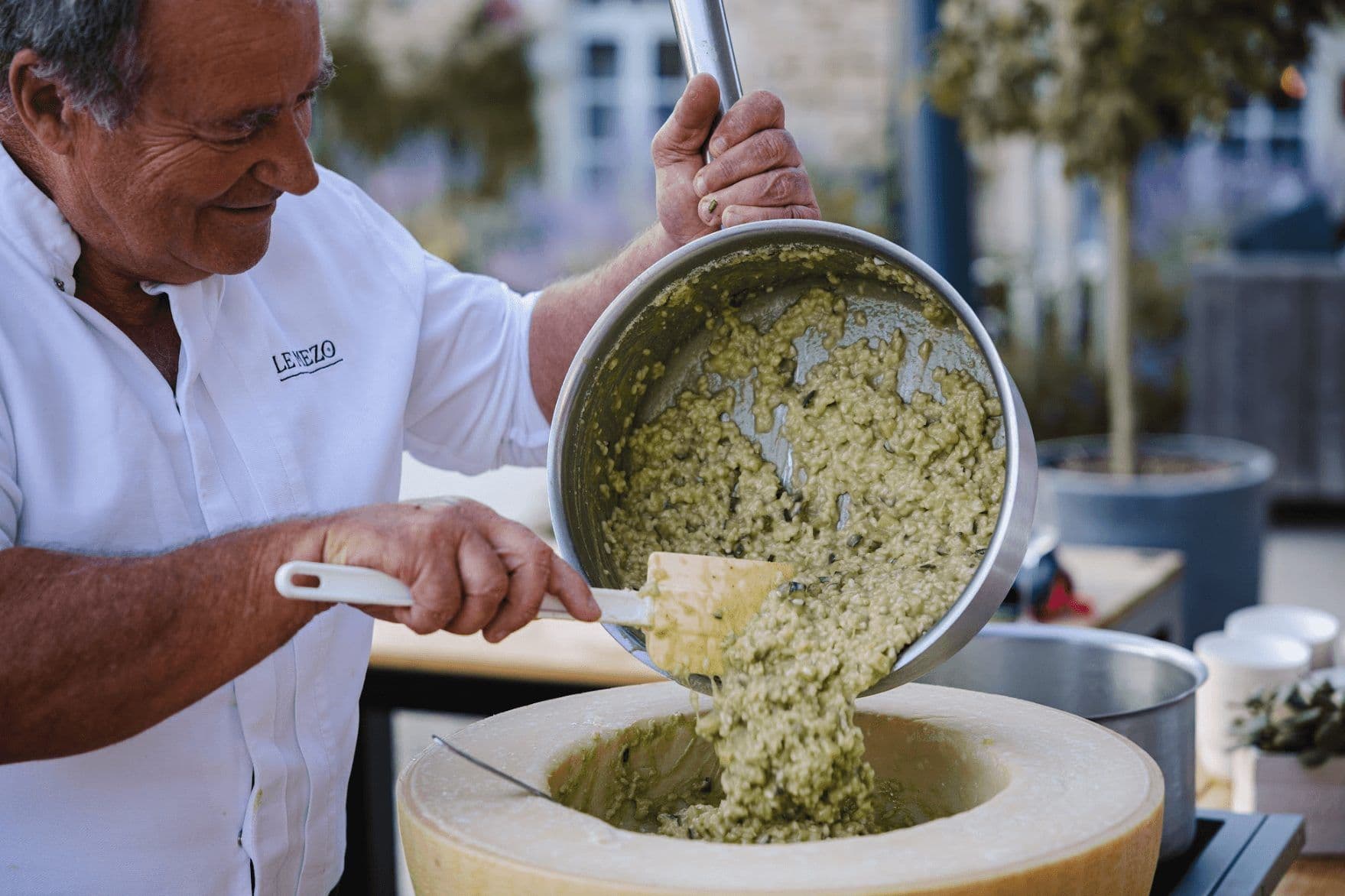Smiling chef pours creamy risotto from a pot into a large cheese wheel, ideal for corporate dinner catering.