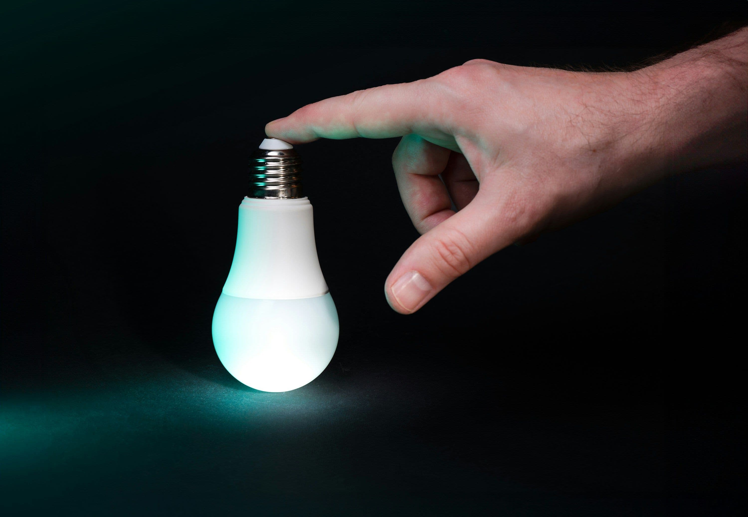 A hand touches a glowing teal LED light bulb on a dark background, symbolizing innovation.