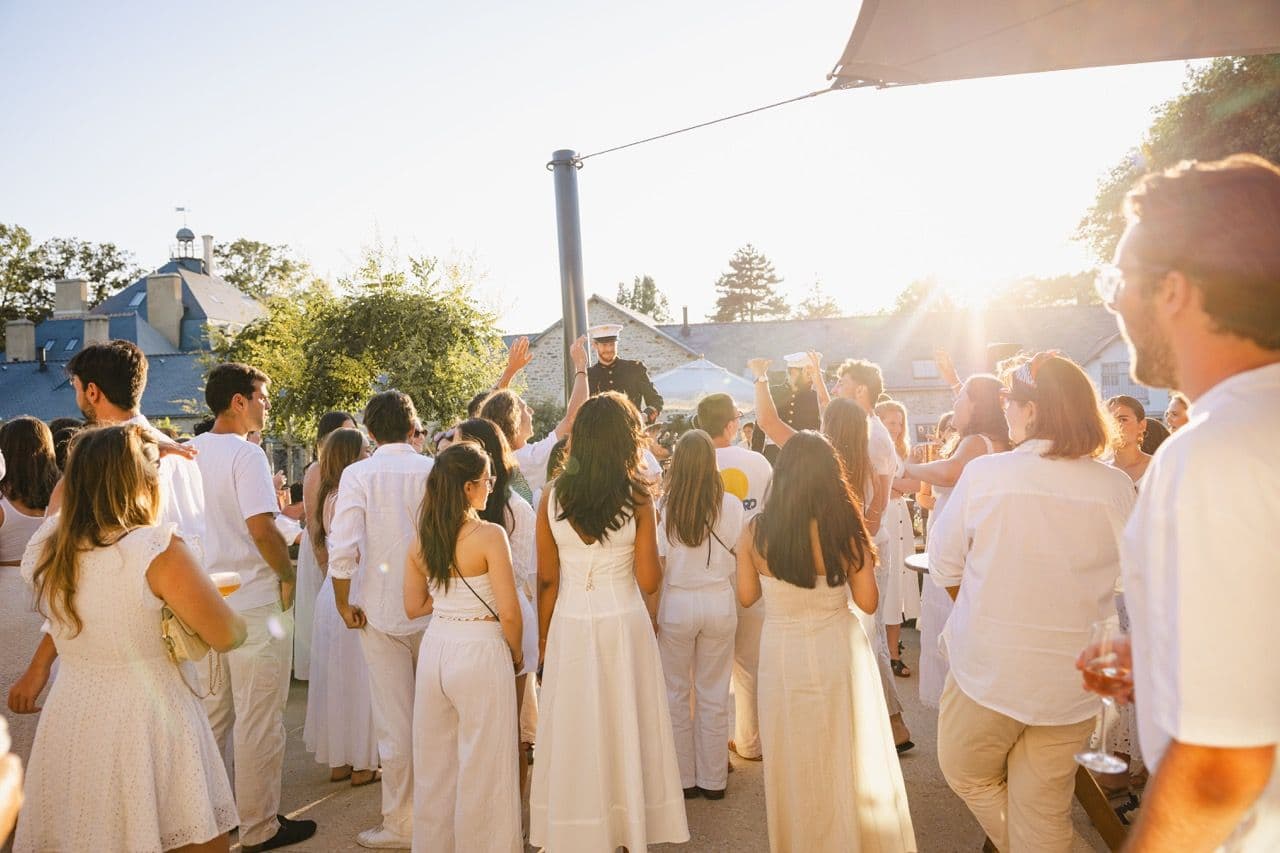 Attendees in white attire celebrate an outdoor corporate event at a historic venue during sunset.