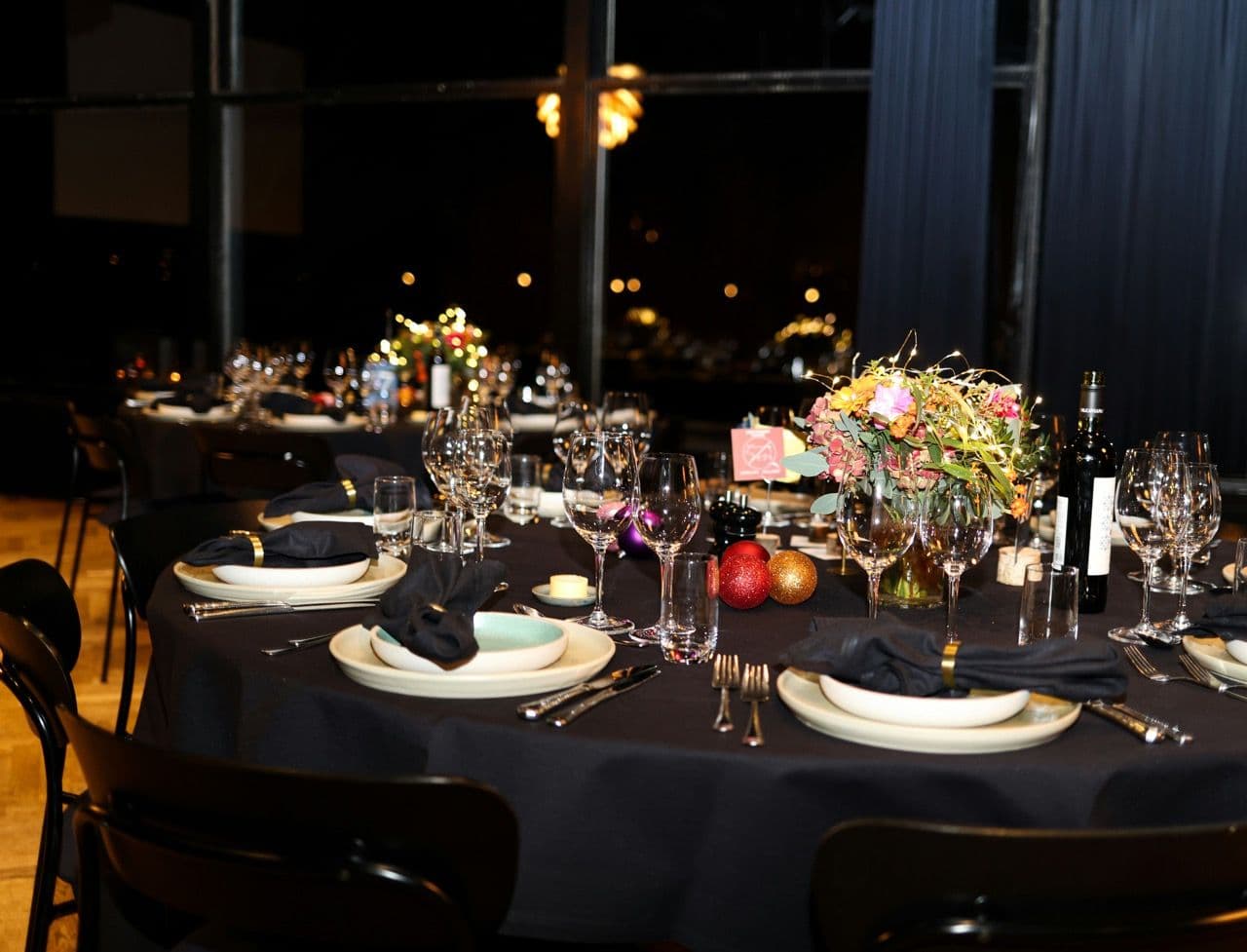 Elegant corporate dinner table setting with black tablecloths, floral centerpiece, and city lights at night.