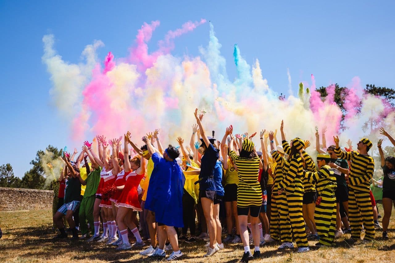 A large group celebrates an outdoor corporate team building event with colorful powder under a blue sky.