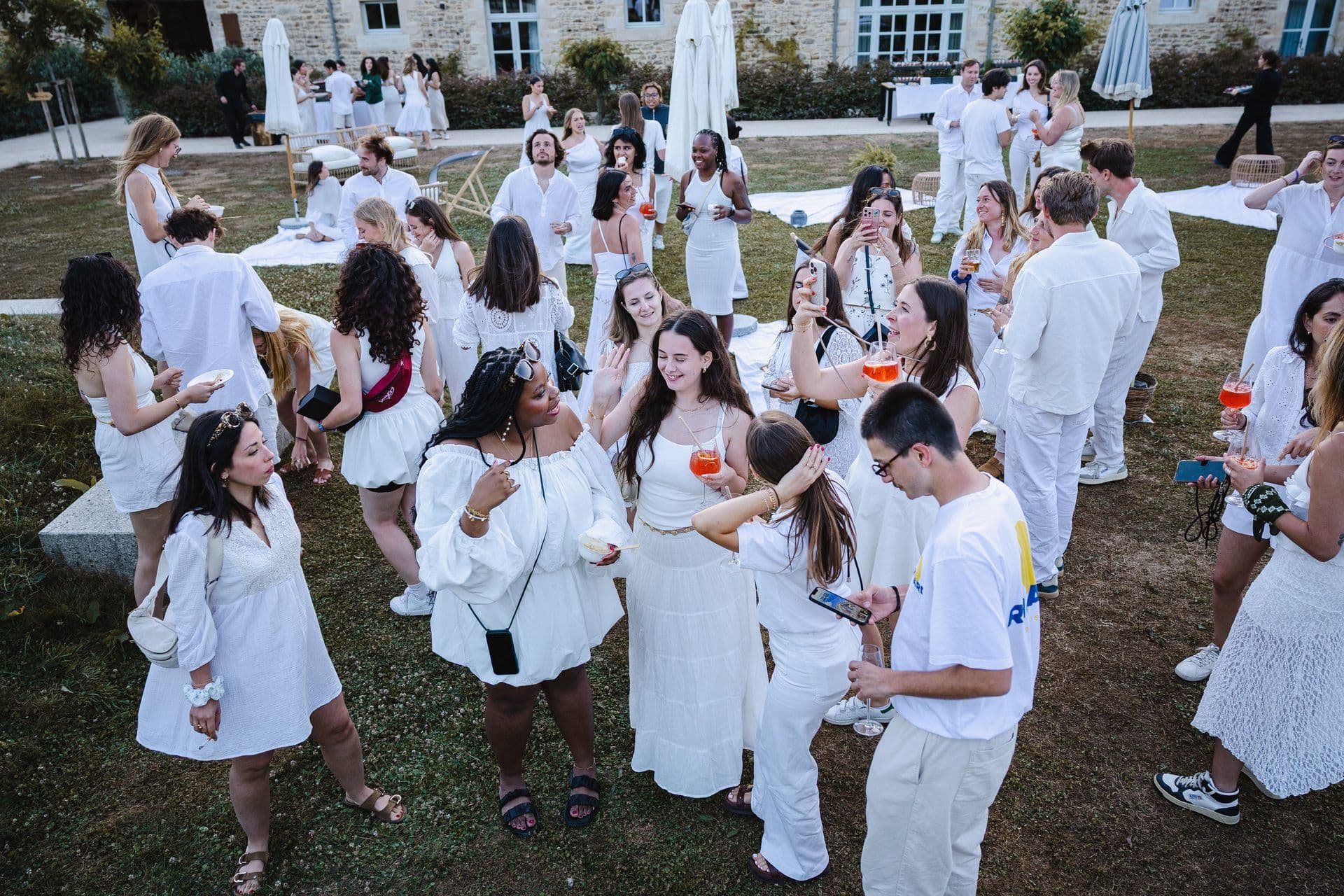 A large group of people in white attire enjoy a corporate event on a grassy lawn outside a historic building.