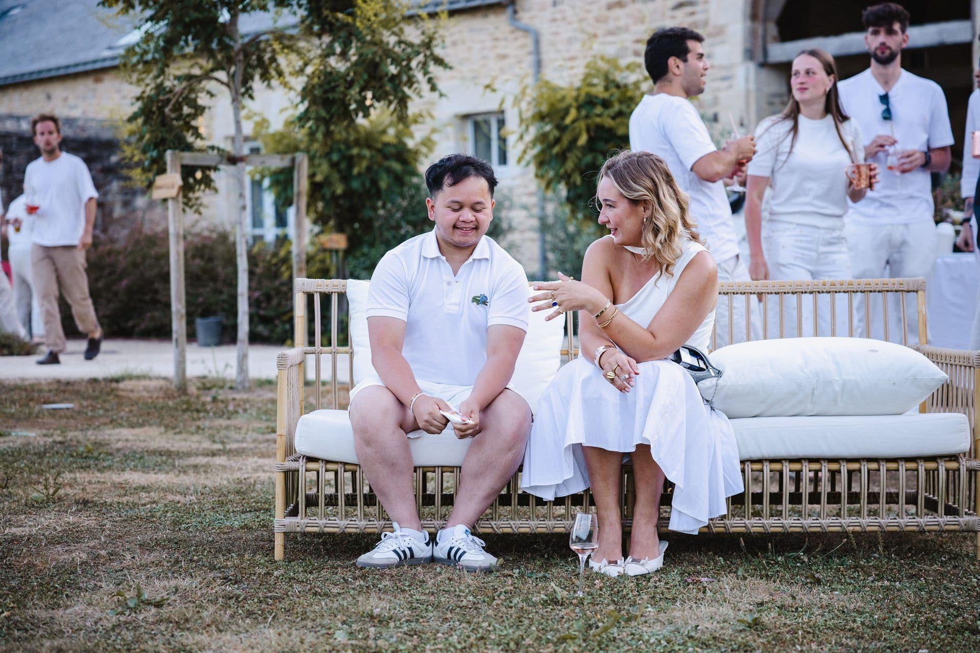 Two colleagues in white attire chat on an outdoor rattan sofa during a corporate offsite event at a historic