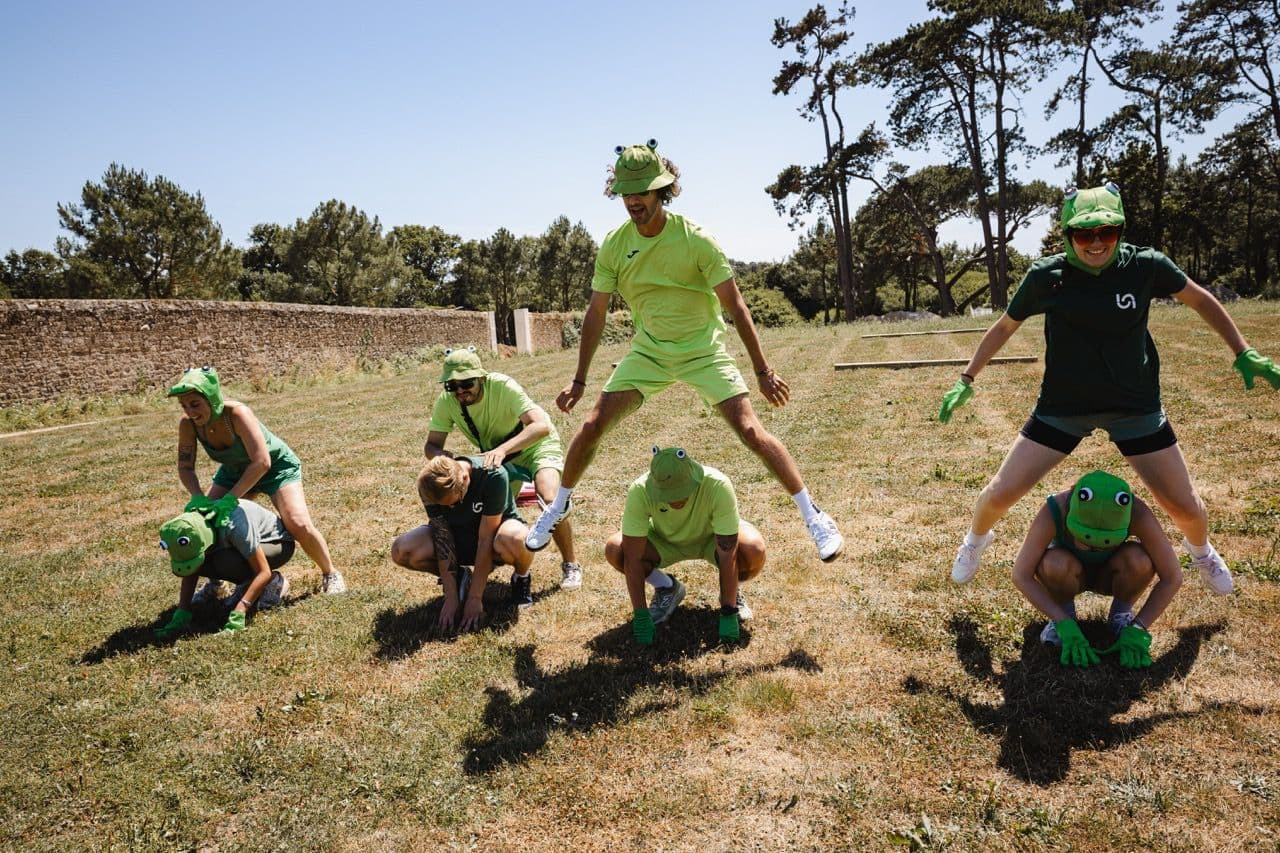A group of colleagues in frog costumes participates in an outdoor team building activity on a sunny day.