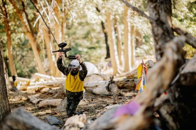 A person in a yellow vest and protective mask kneels, holding a paintball gun during an outdoor team activity.