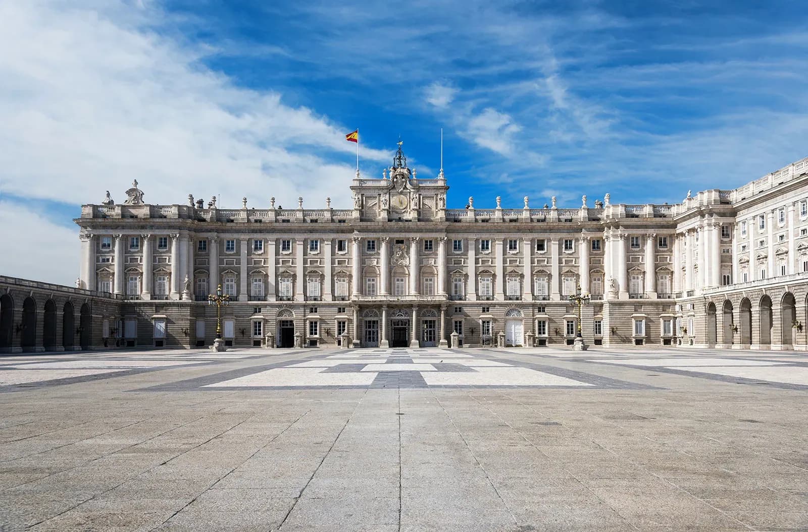 Grand Royal Palace of Madrid, Spain, with a Spanish flag and clock tower, overlooking a large plaza.