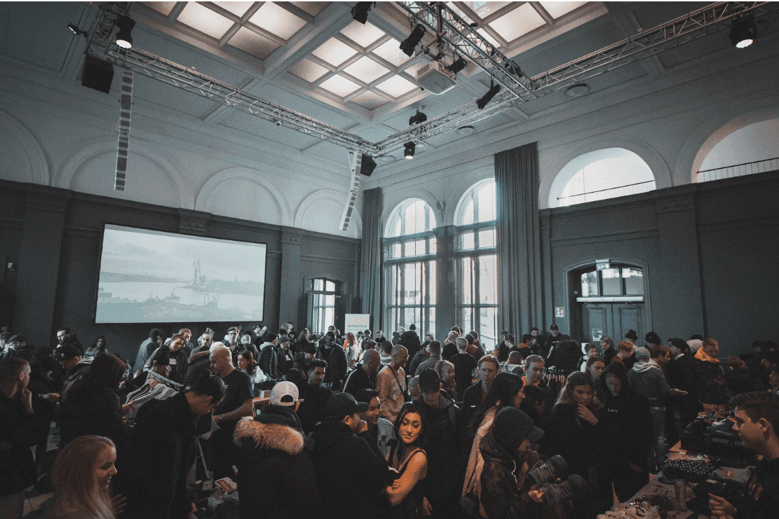 A large crowd of attendees gathers in a spacious corporate event venue with a high ceiling, arched windows