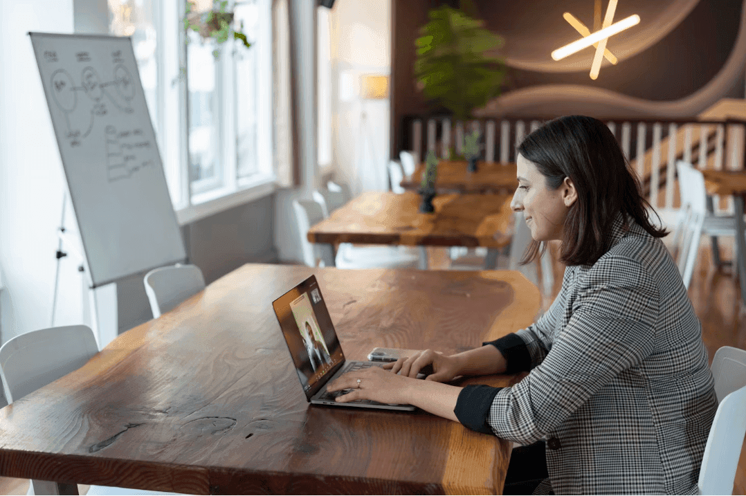 Woman on a video call at a large wooden table in a modern corporate event venue with a whiteboard.