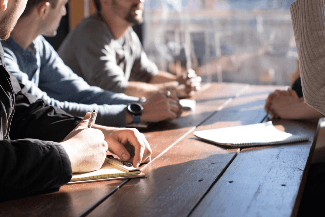 Professionals take notes at a wooden table during a corporate seminar or workshop in a bright meeting room.