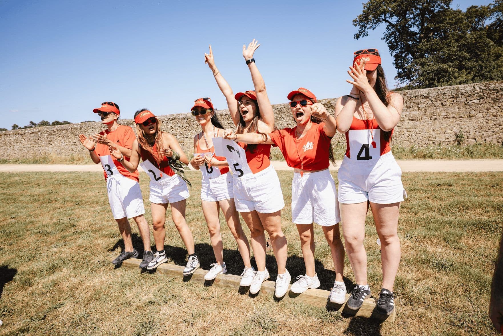 Six colleagues in red and white cheering enthusiastically during an outdoor team building activity.