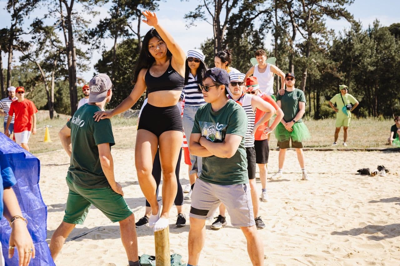 Team members balance on a pole during an outdoor team building activity on a sunny beach.