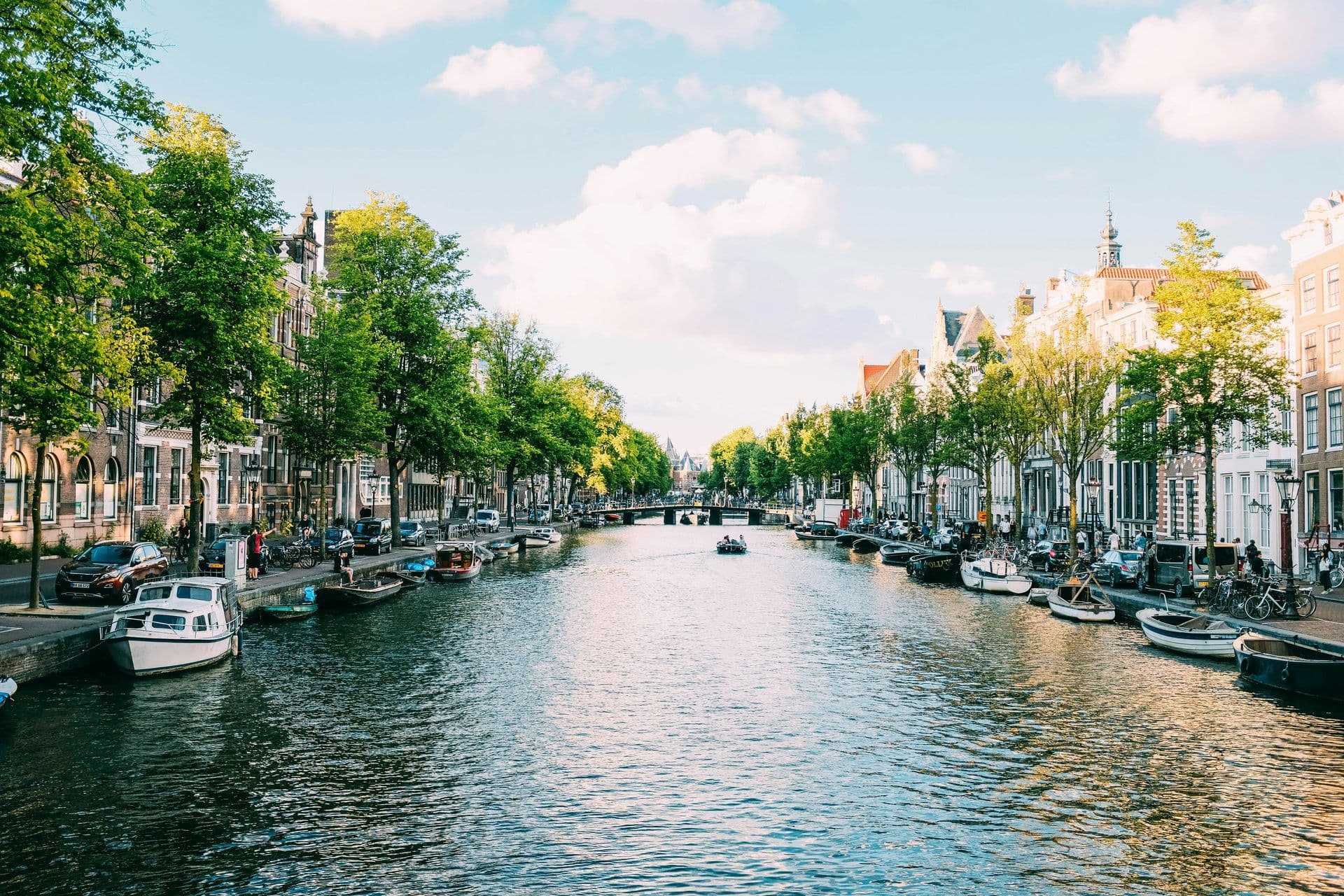 A picturesque canal in Amsterdam lined with historic buildings, green trees, and boats, under a blue sky.
