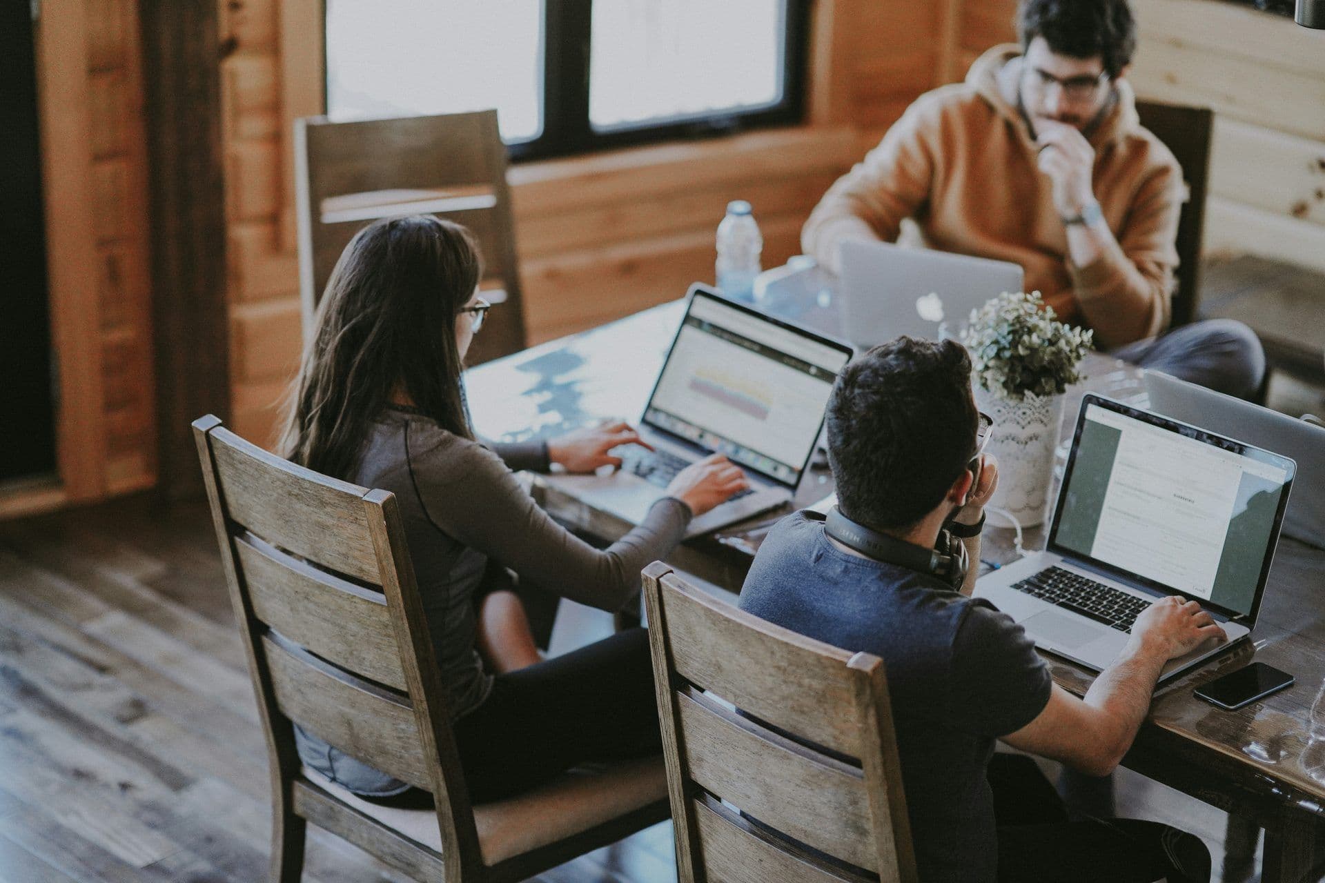 Three colleagues work on laptops at a rustic wooden table during a team offsite or corporate workshop.