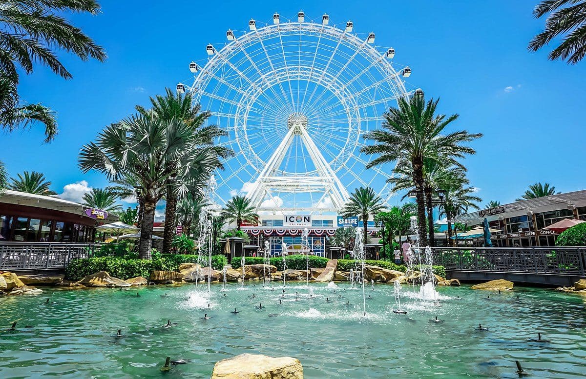 The Orlando Eye Ferris wheel towers over a vibrant outdoor event space with fountains and palm trees in