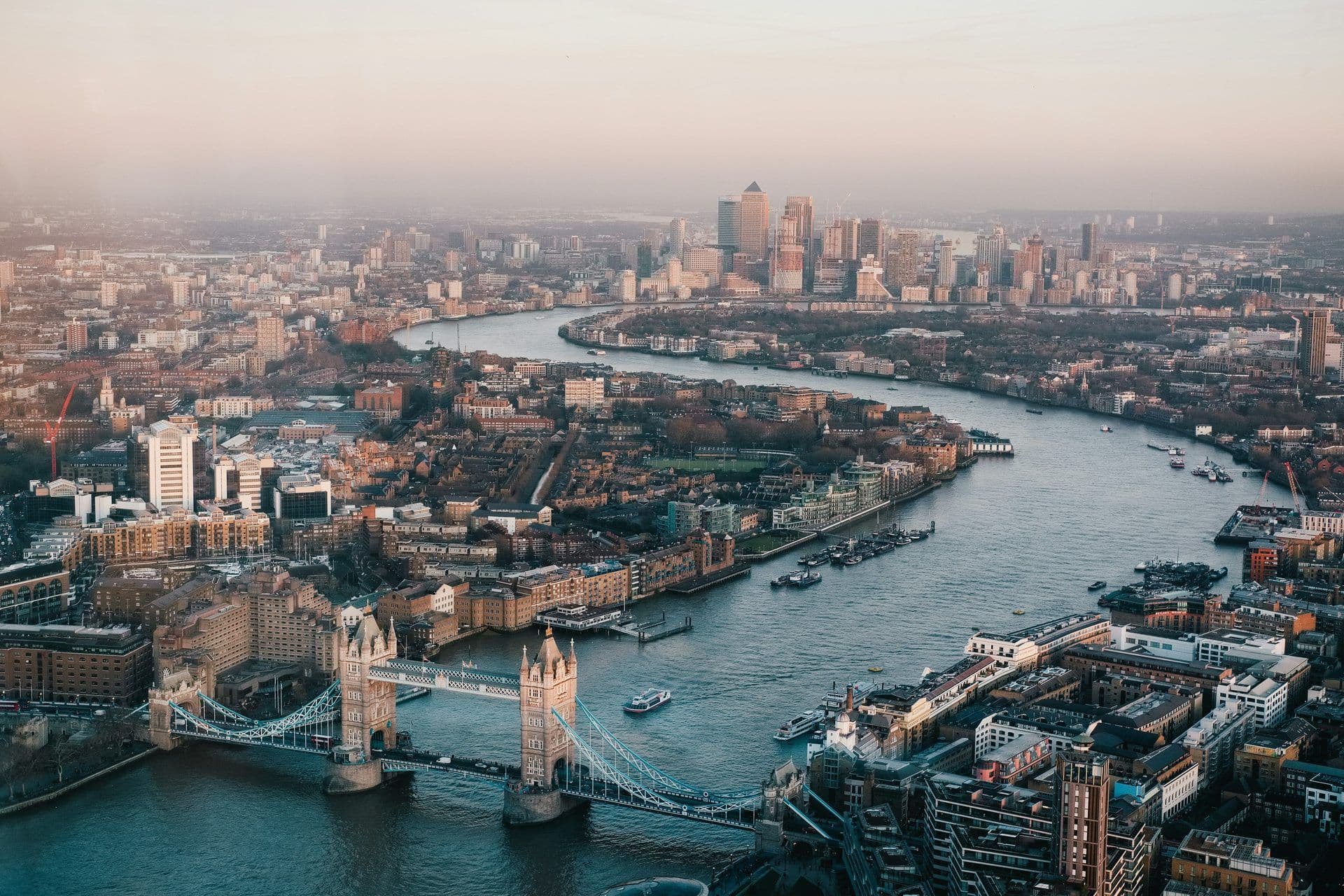 Aerial view of London's River Thames, Tower Bridge, and Canary Wharf skyline at dusk.