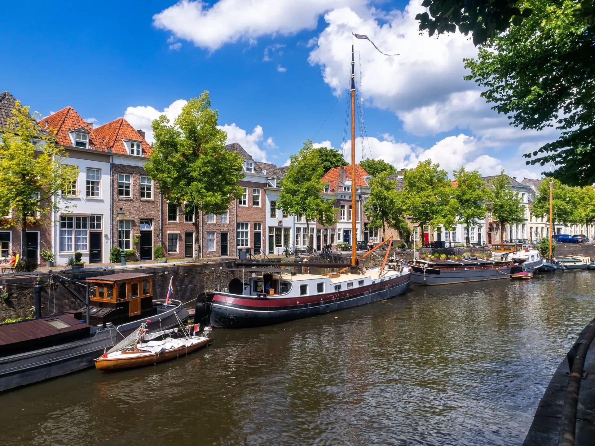 Historic canal in 's-Hertogenbosch, Netherlands, with traditional buildings, green trees, and moored boats.