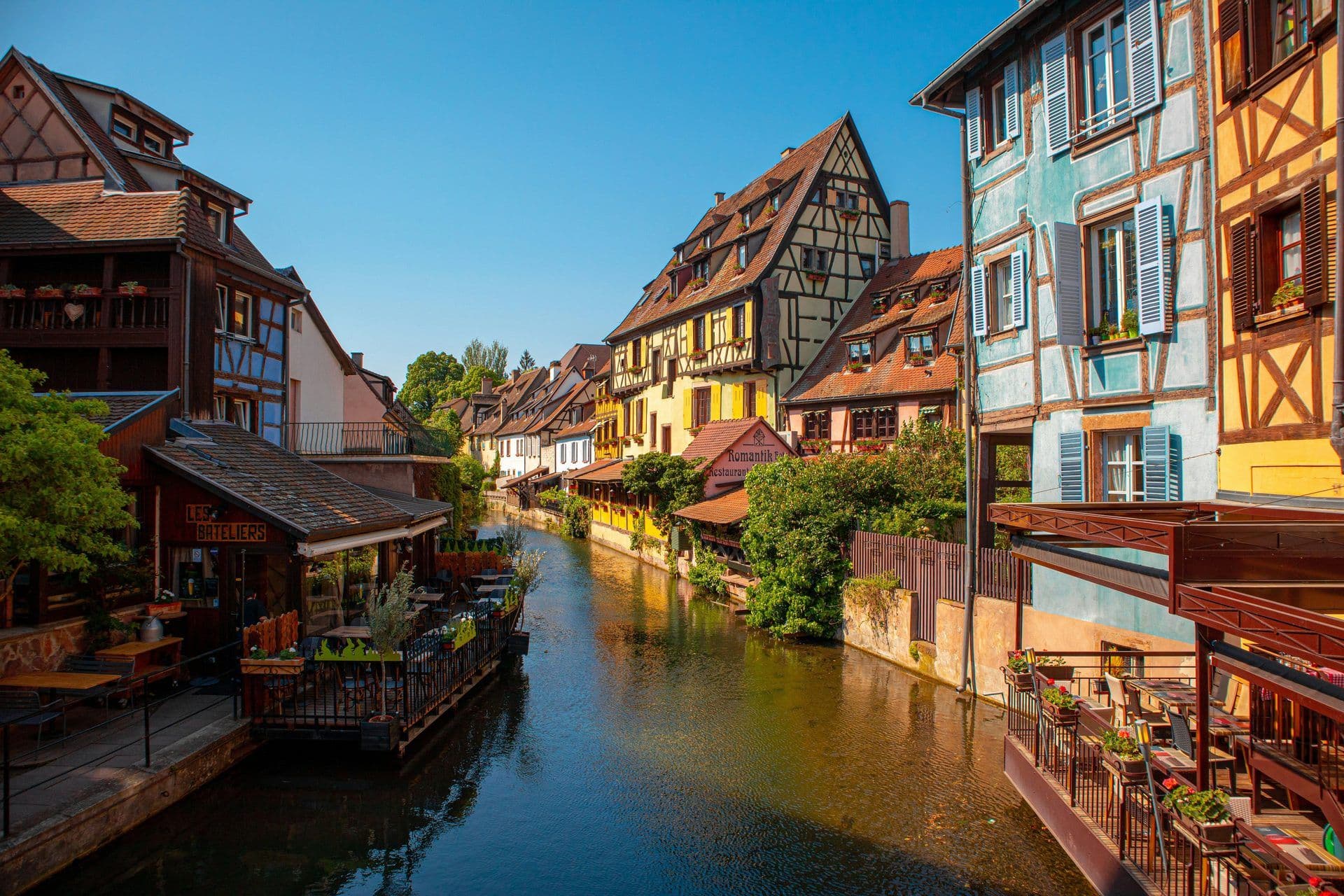 A scenic canal in Colmar, France, lined with colorful half-timbered buildings and outdoor restaurant terraces.