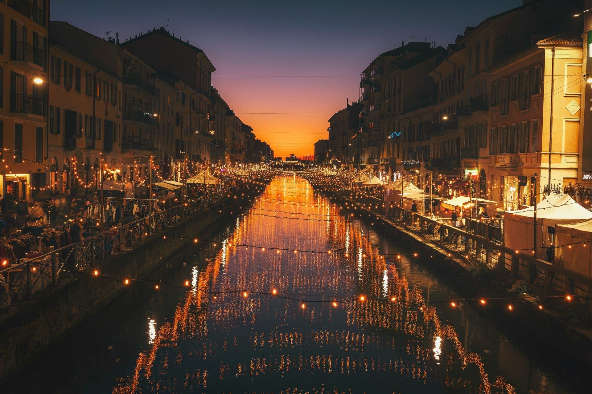 Historic Milan canal at dusk, featuring a lively outdoor event space with string lights and market stalls.
