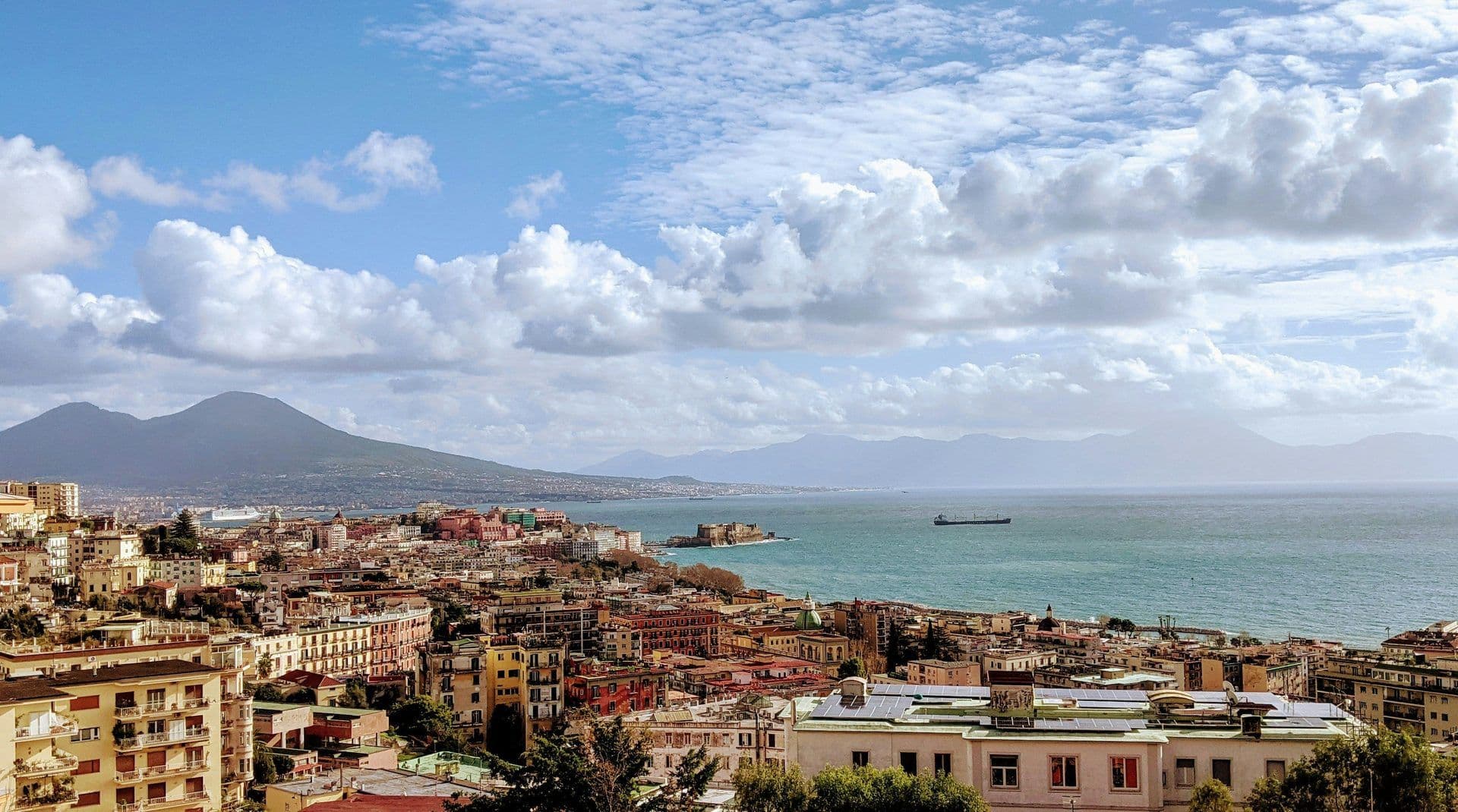 Panoramic view of Naples, Italy, featuring colorful buildings, a bay, and Mount Vesuvius.