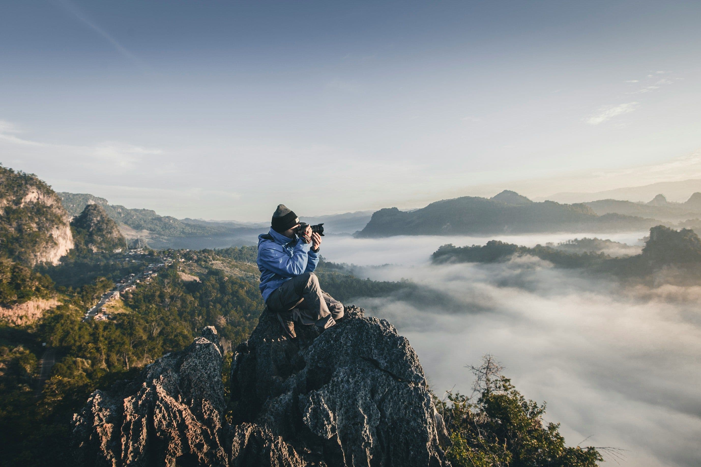 An event photographer in a blue jacket shooting from a mountain peak above the clouds at sunrise