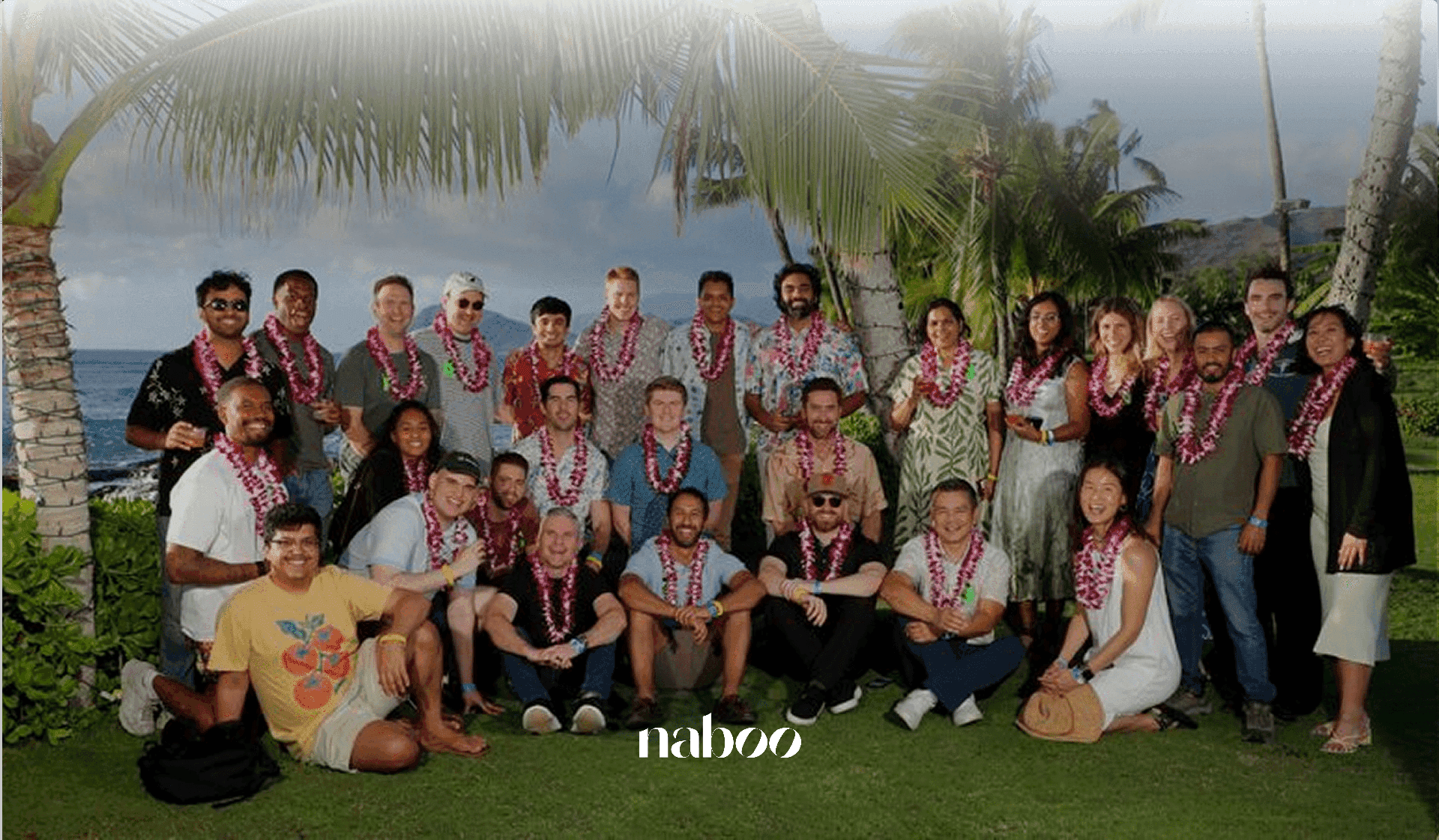 A large corporate team wearing leis poses on a grassy outdoor waterfront venue for a team offsite.