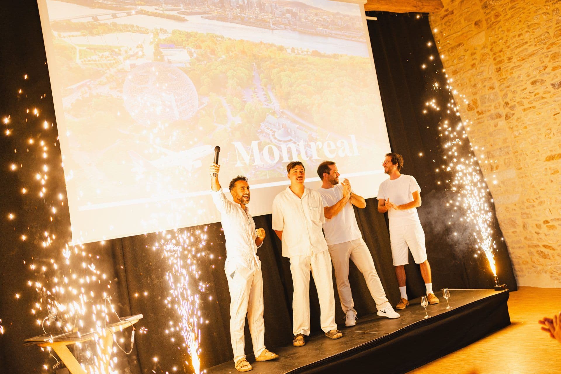 Four men celebrate on stage with indoor fireworks at a corporate event, screen shows Montreal.