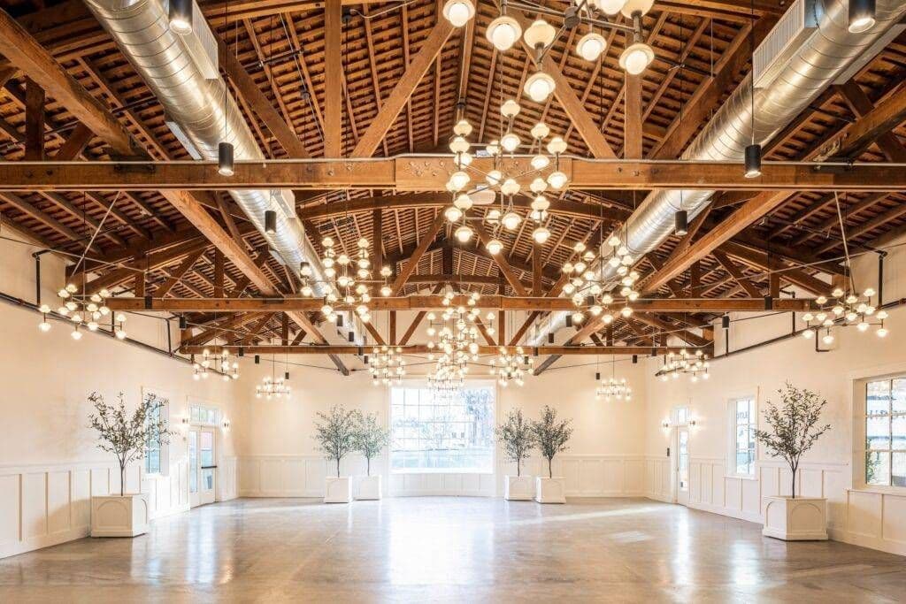 Modern industrial wedding venue with exposed wooden beams, chandeliers, and polished wooden floor.