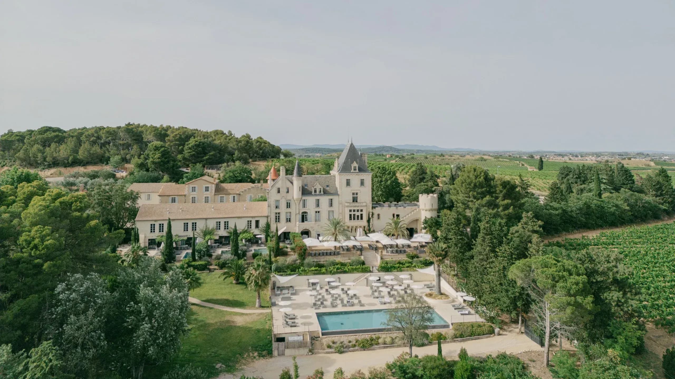 Aerial view of a grand château with towers, pool, and manicured gardens surrounded by vineyards and forests in Provence.