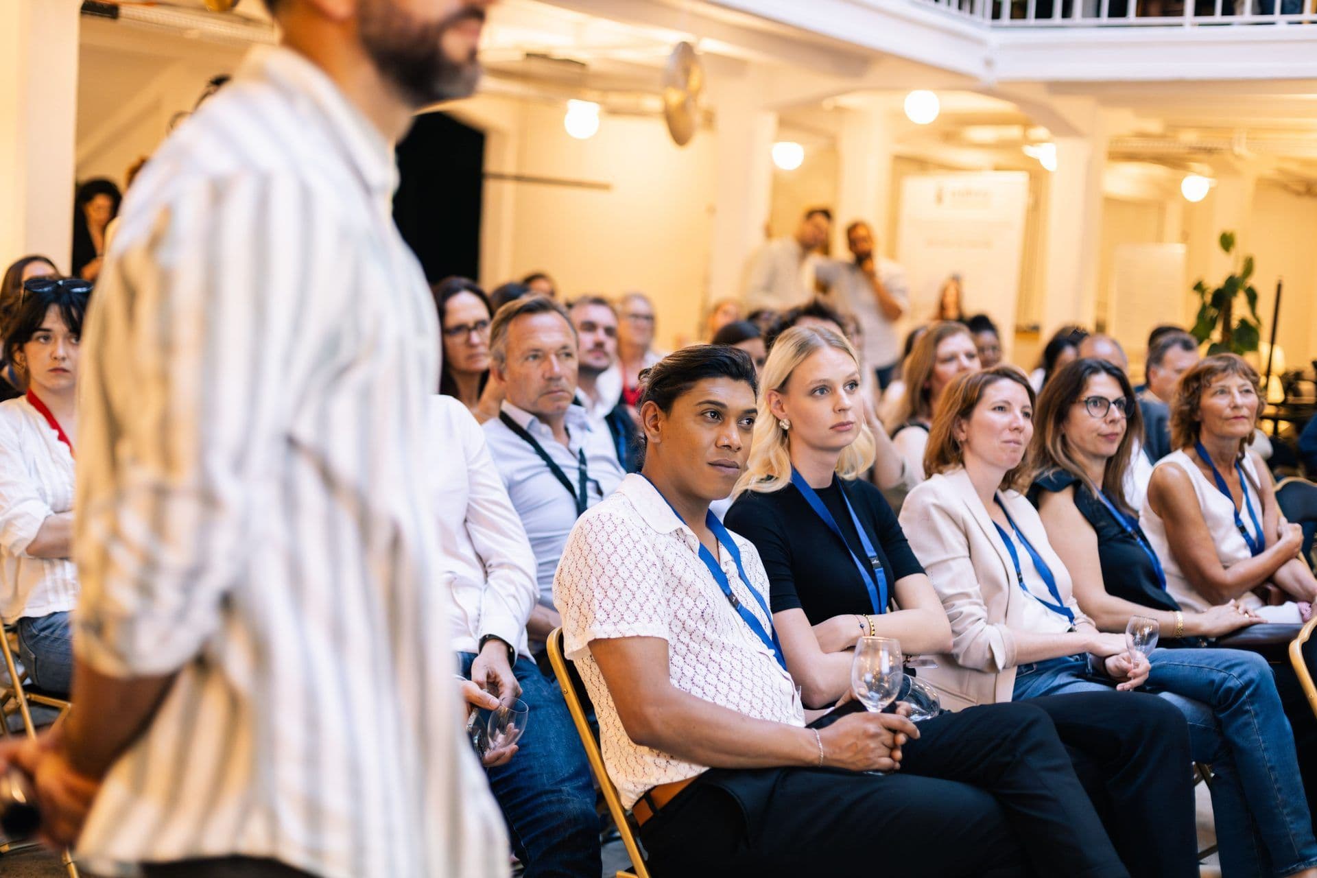 Audience members wearing blue lanyards listen attentively to a speaker at a professional conference event in a well-lit indoo