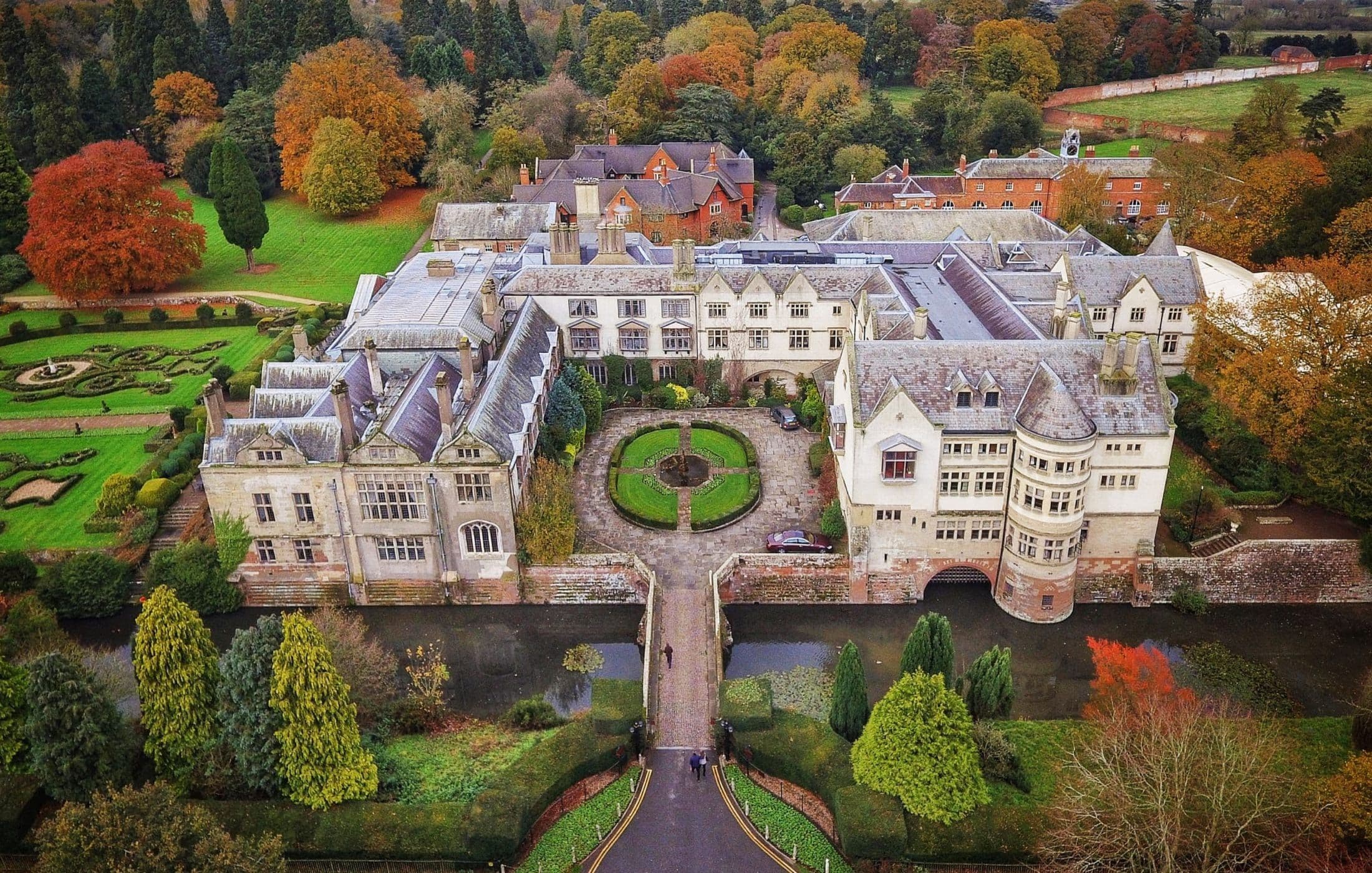 Aerial view of a grand English castle with manicured gardens, surrounded by moat and autumn woodland.