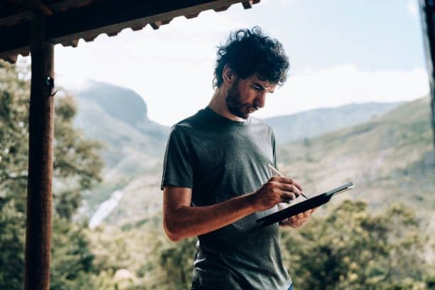 Curly-haired man using a tablet and stylus outdoors, with a scenic mountain and forest backdrop.