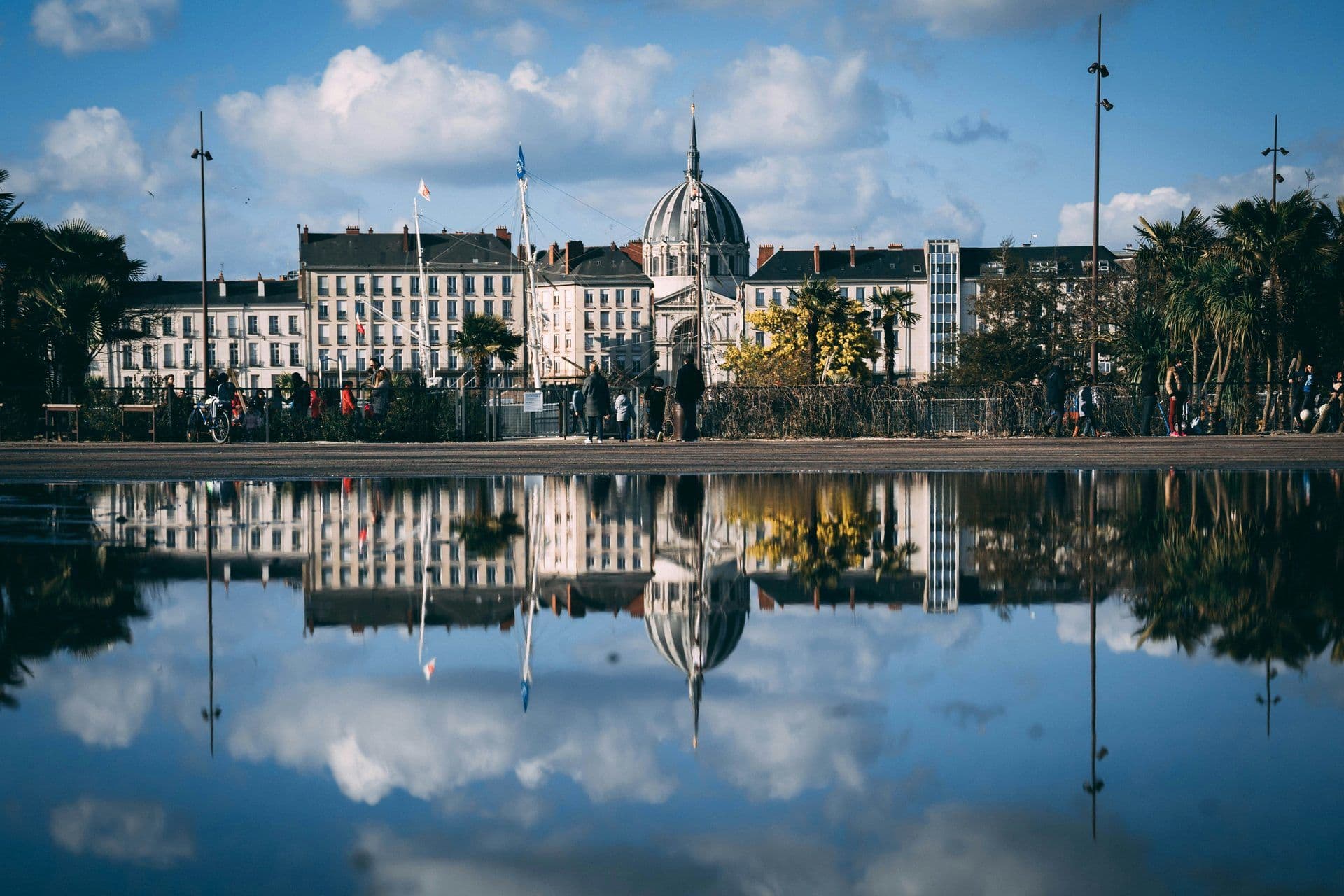 A historic domed building and city architecture reflect in a large puddle in La Rochelle, France.