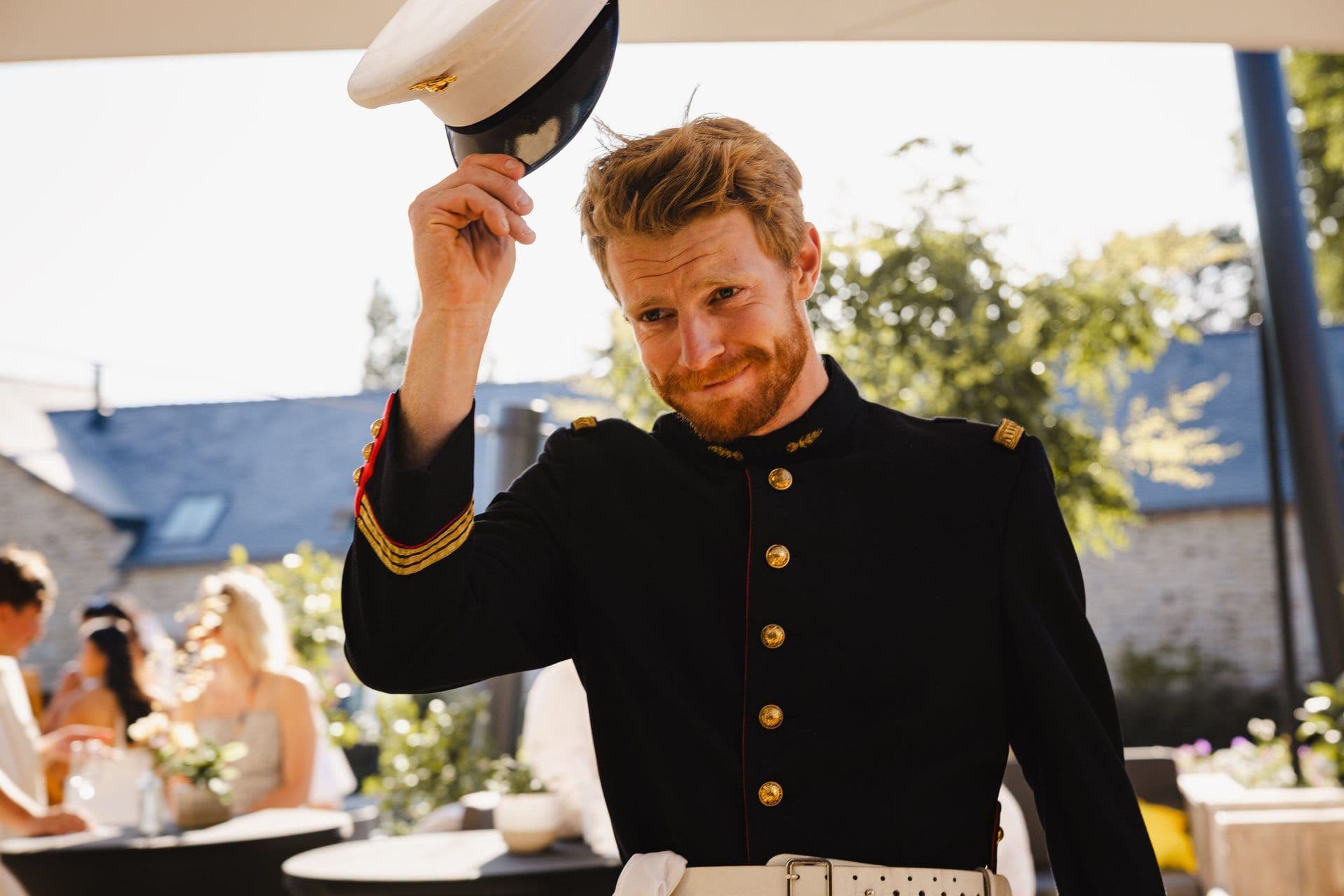 A smiling man in a dark uniform with gold trim holds his cap at an outdoor corporate event venue.