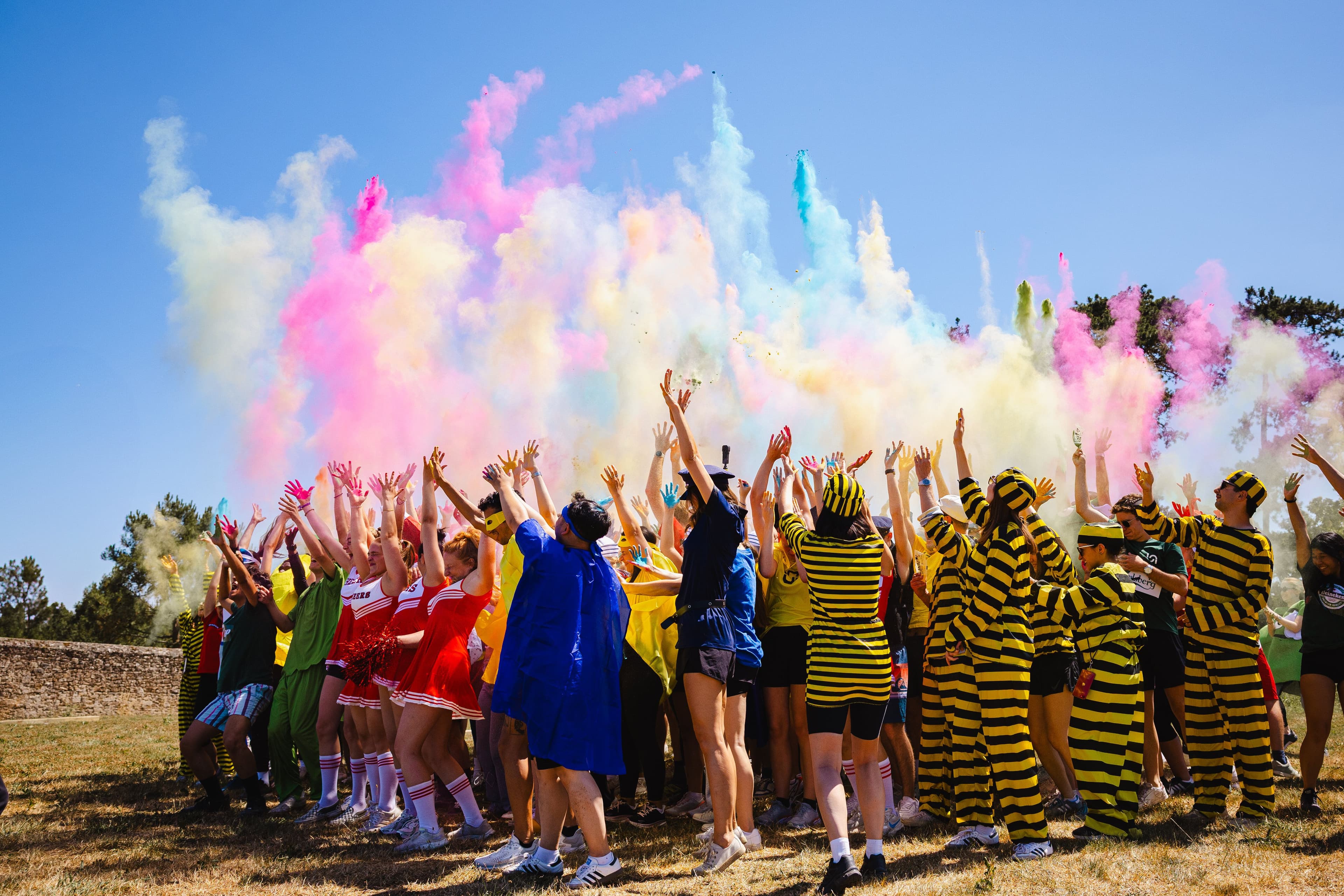 Employees in costumes raise their arms as colorful powder explodes during an outdoor team building event.