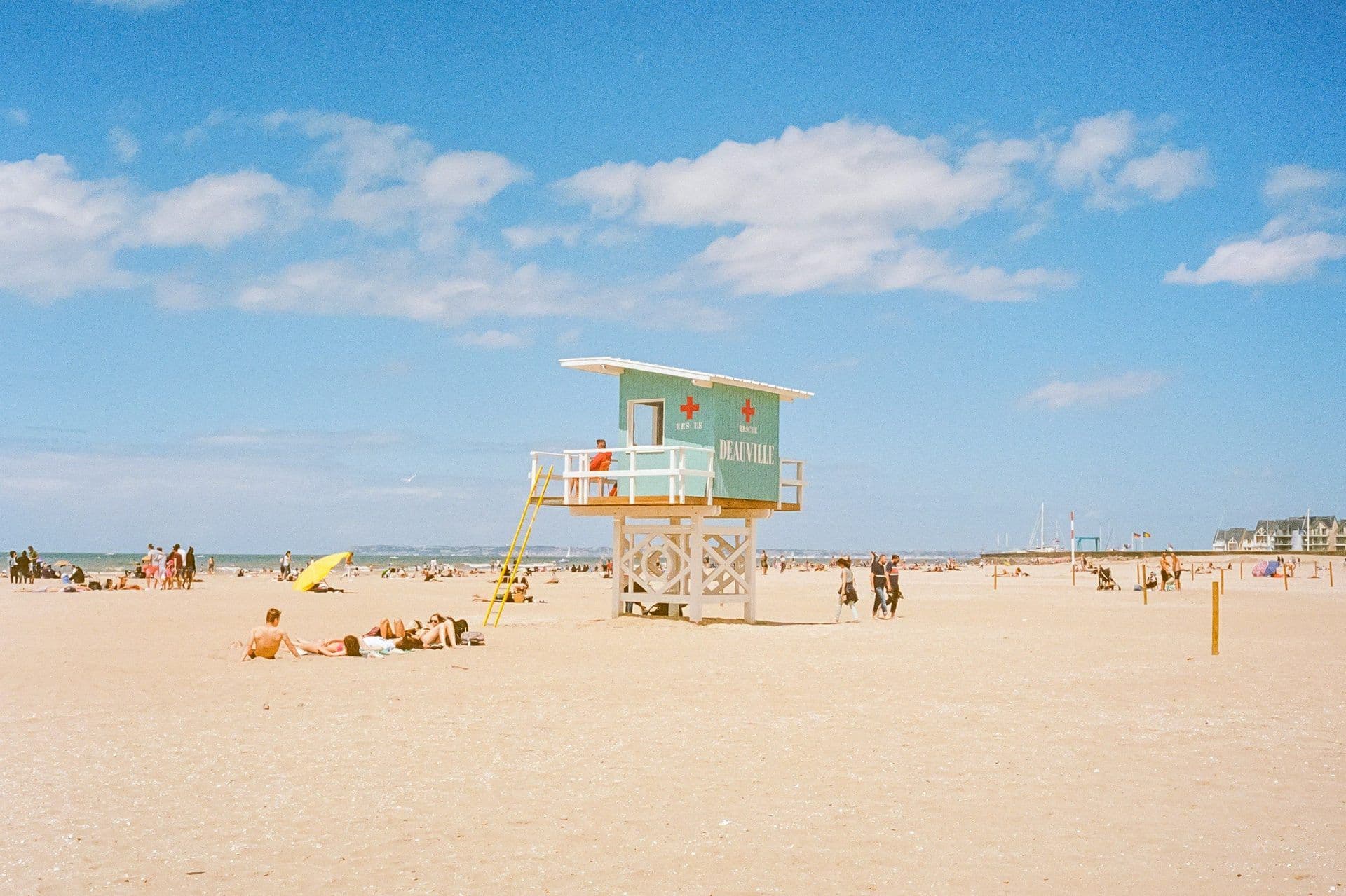 A turquoise lifeguard station stands on a sunny sandy beach in Deauville, France, with people relaxing.