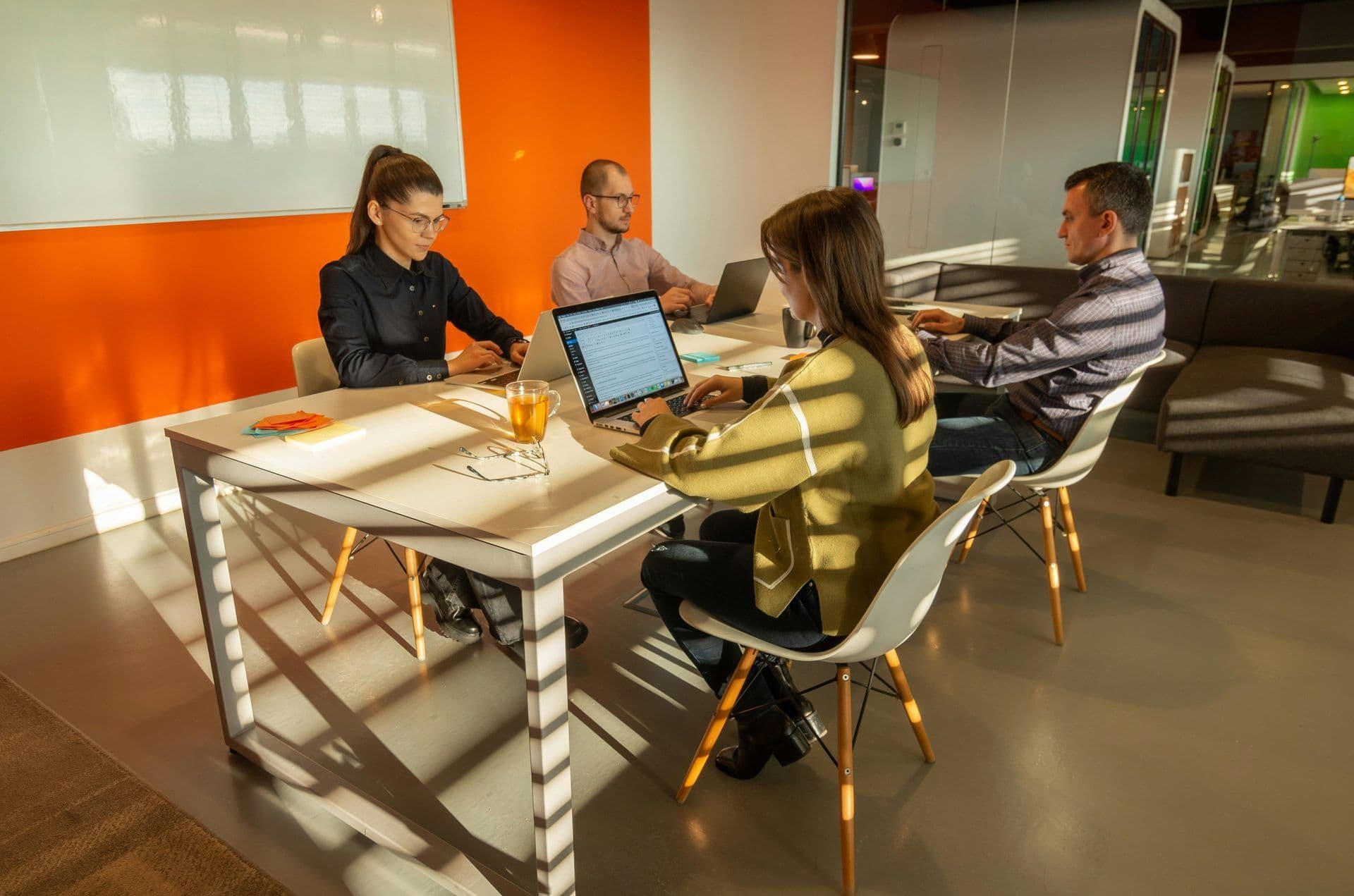 Three colleagues work on laptops at a white table in a modern meeting room with an orange accent wall.