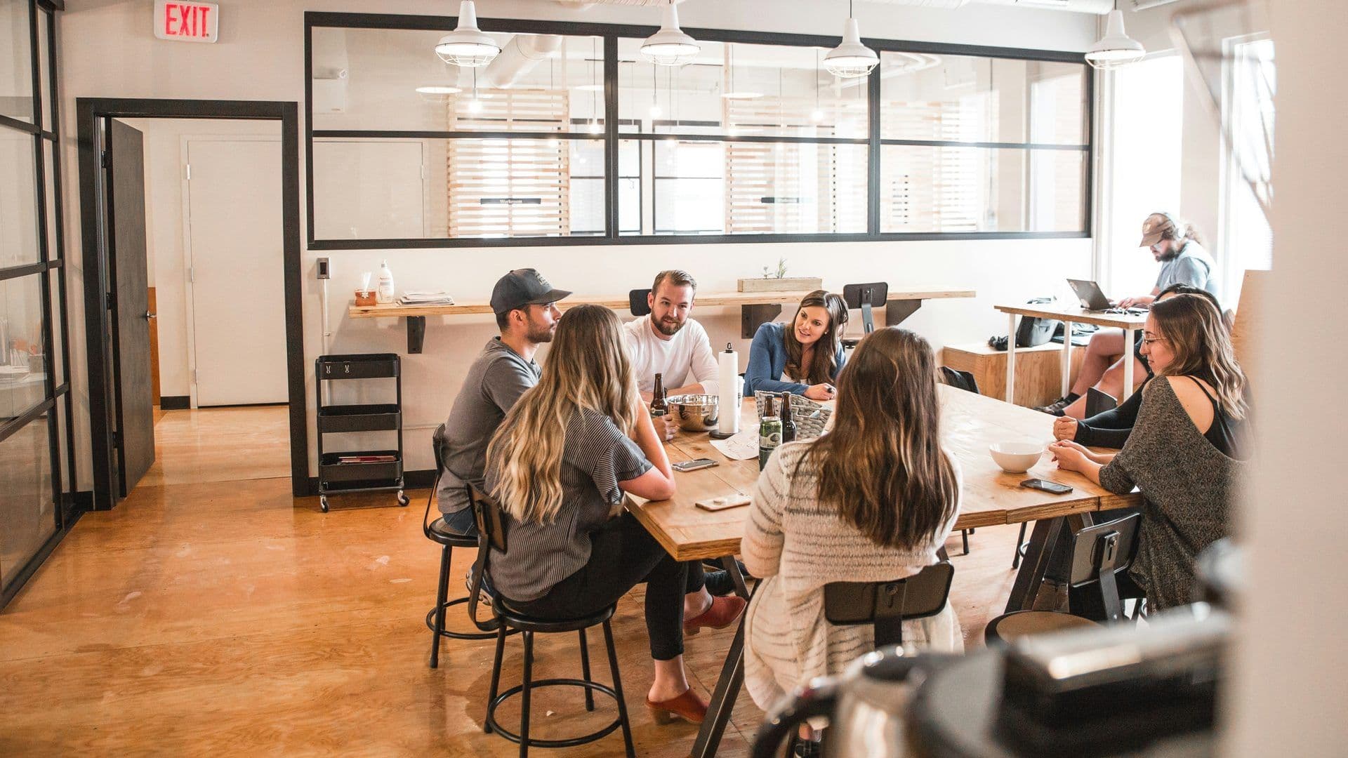 Seven team members discuss ideas at a large wooden table in a contemporary office event space.