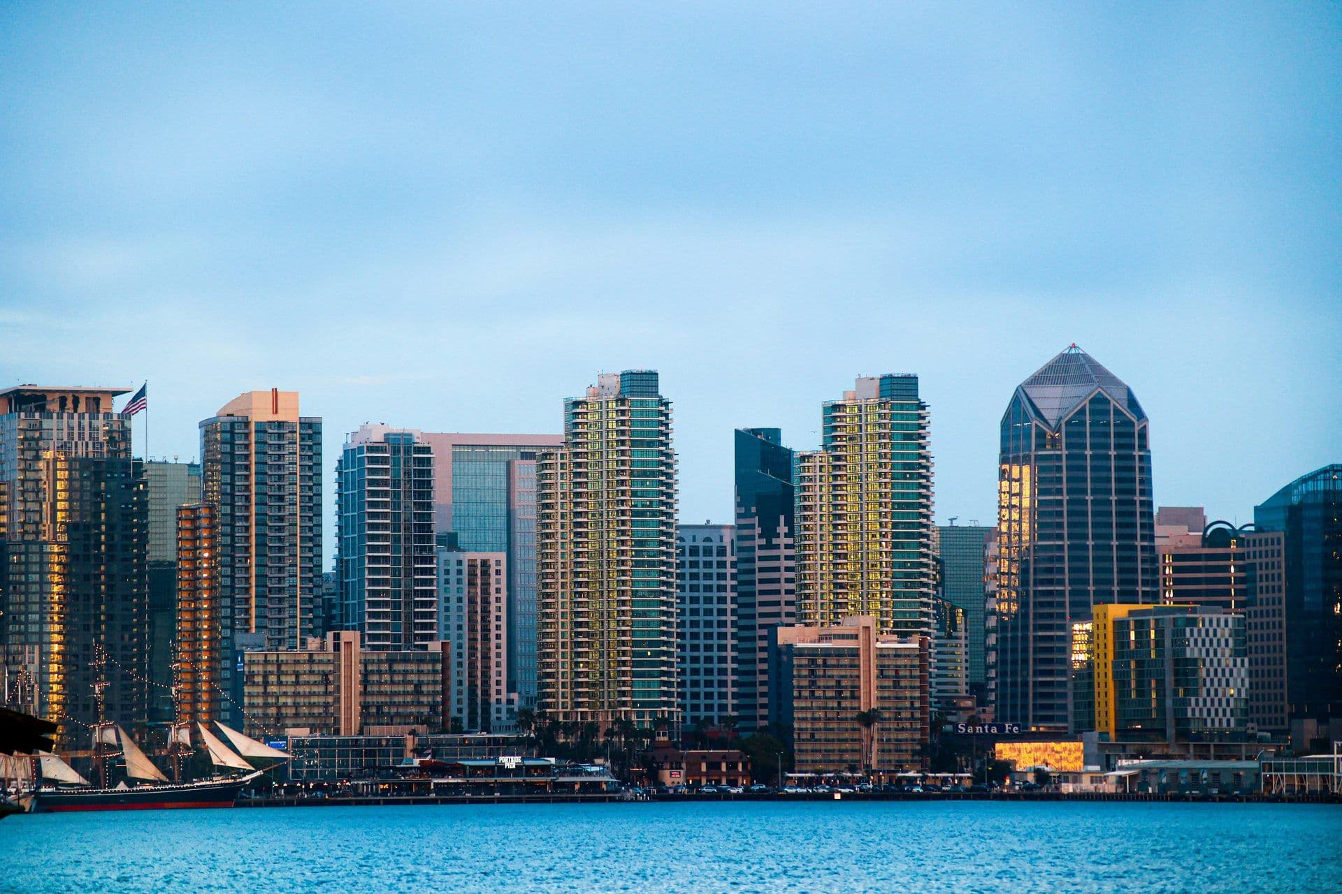 San Diego skyline at dusk with illuminated high-rise buildings, a tall ship, and a waterfront view.