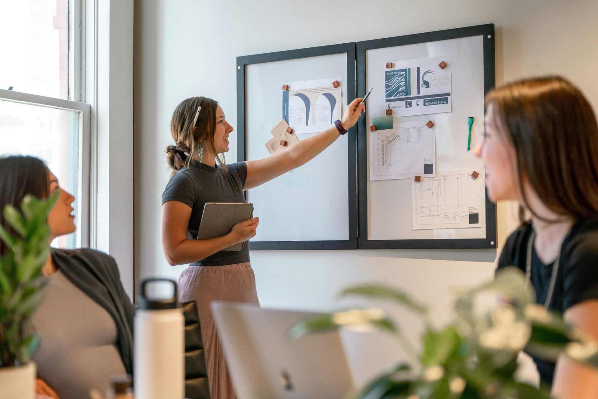 A woman leads a workshop, pointing at a mood board with branding and design concepts in a modern meeting room.